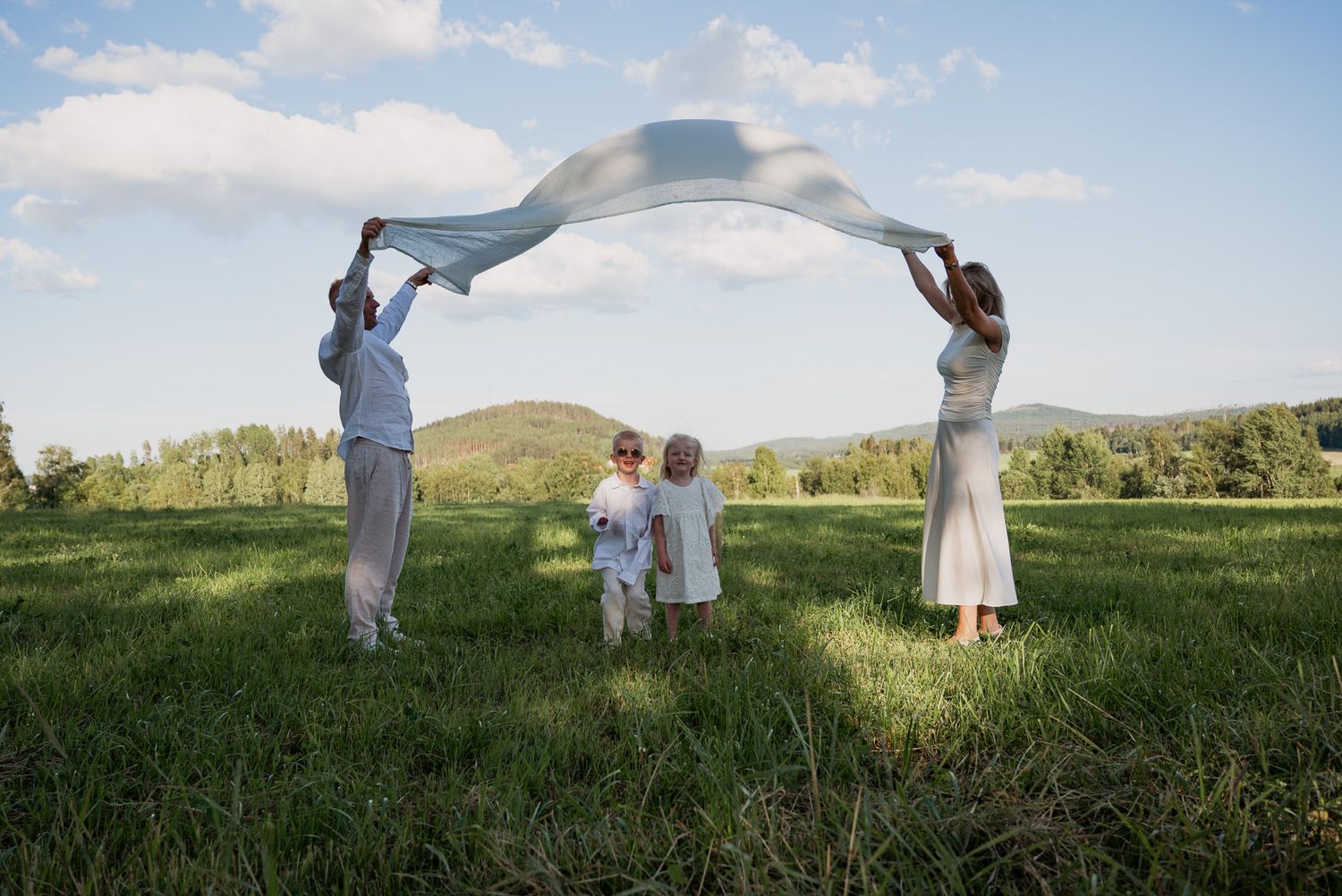 Familj leker på äng med lakan under fotografering på en sommaräng i kvällssolen.