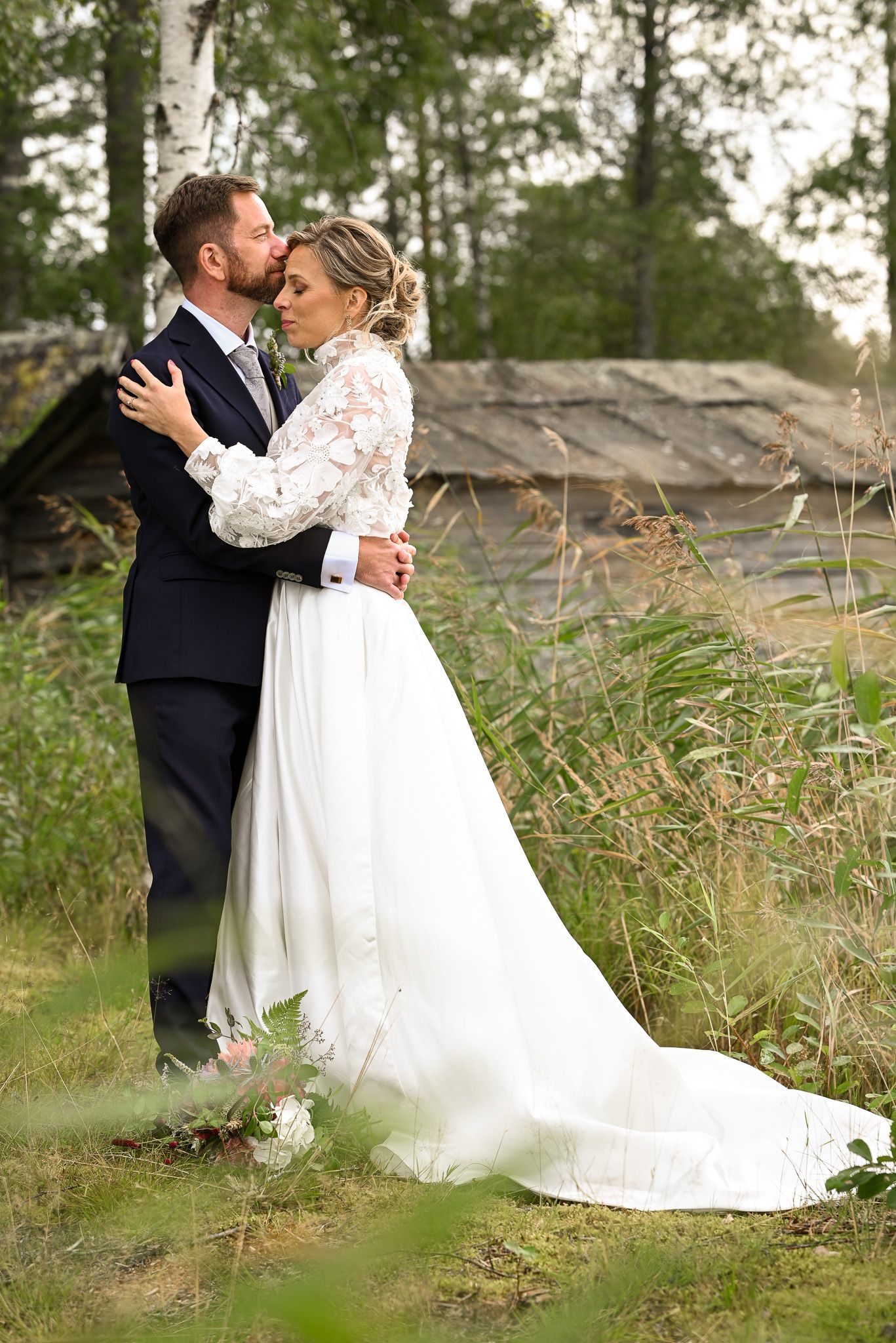 Romantiskt par håller om varandra under bröllopsfoto session i högt gräs intill sjöbodar.