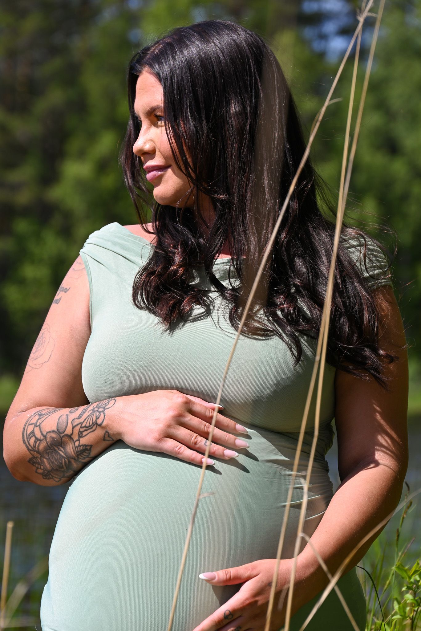 Gravid kvinna i mintgrön klänning ler och håller sig på magen medan hon står i högt gräs intill en sjö.