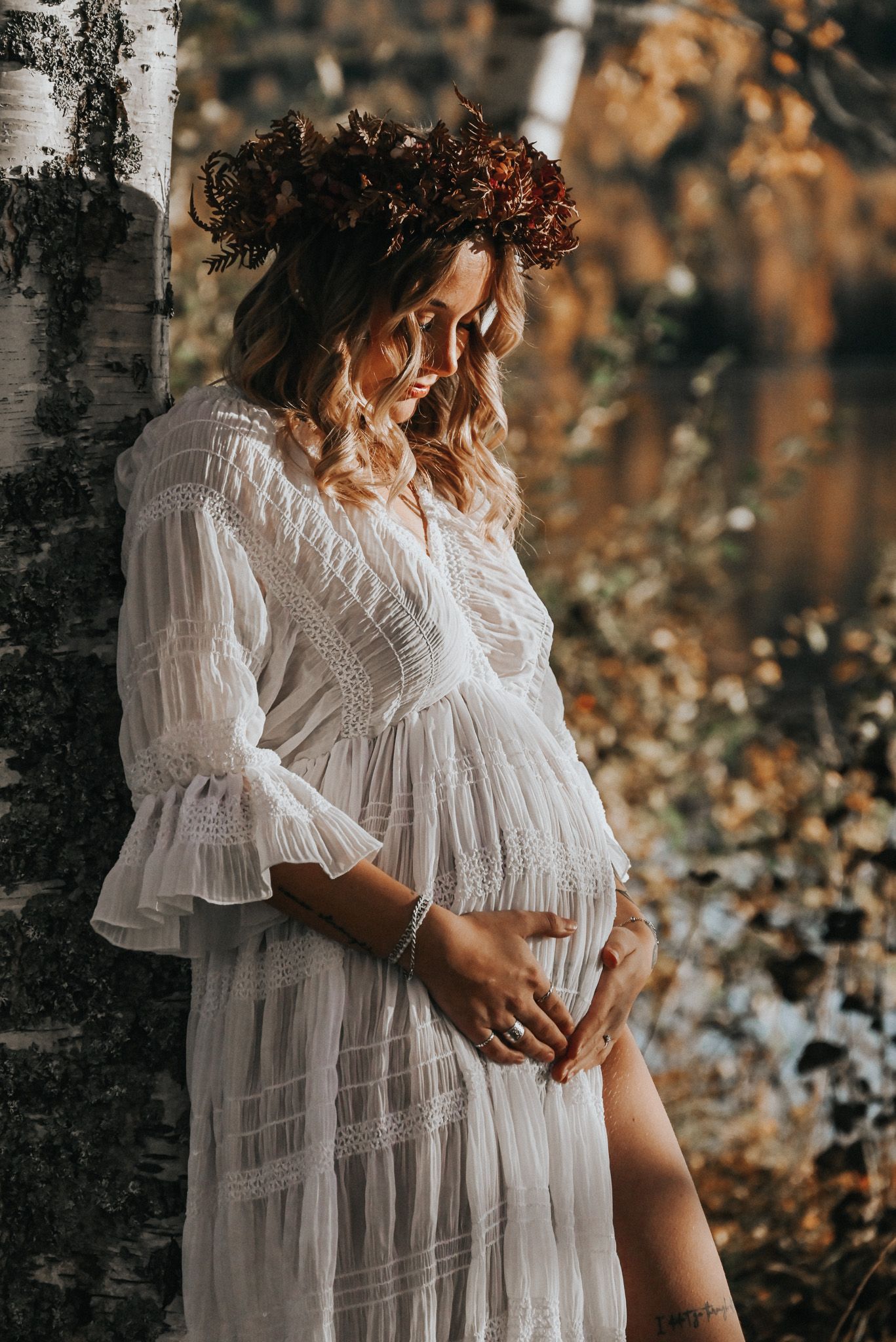 Gravid kvinna poserar i vit spetsklänning med blomkrans i håret och en höstig backdrop.