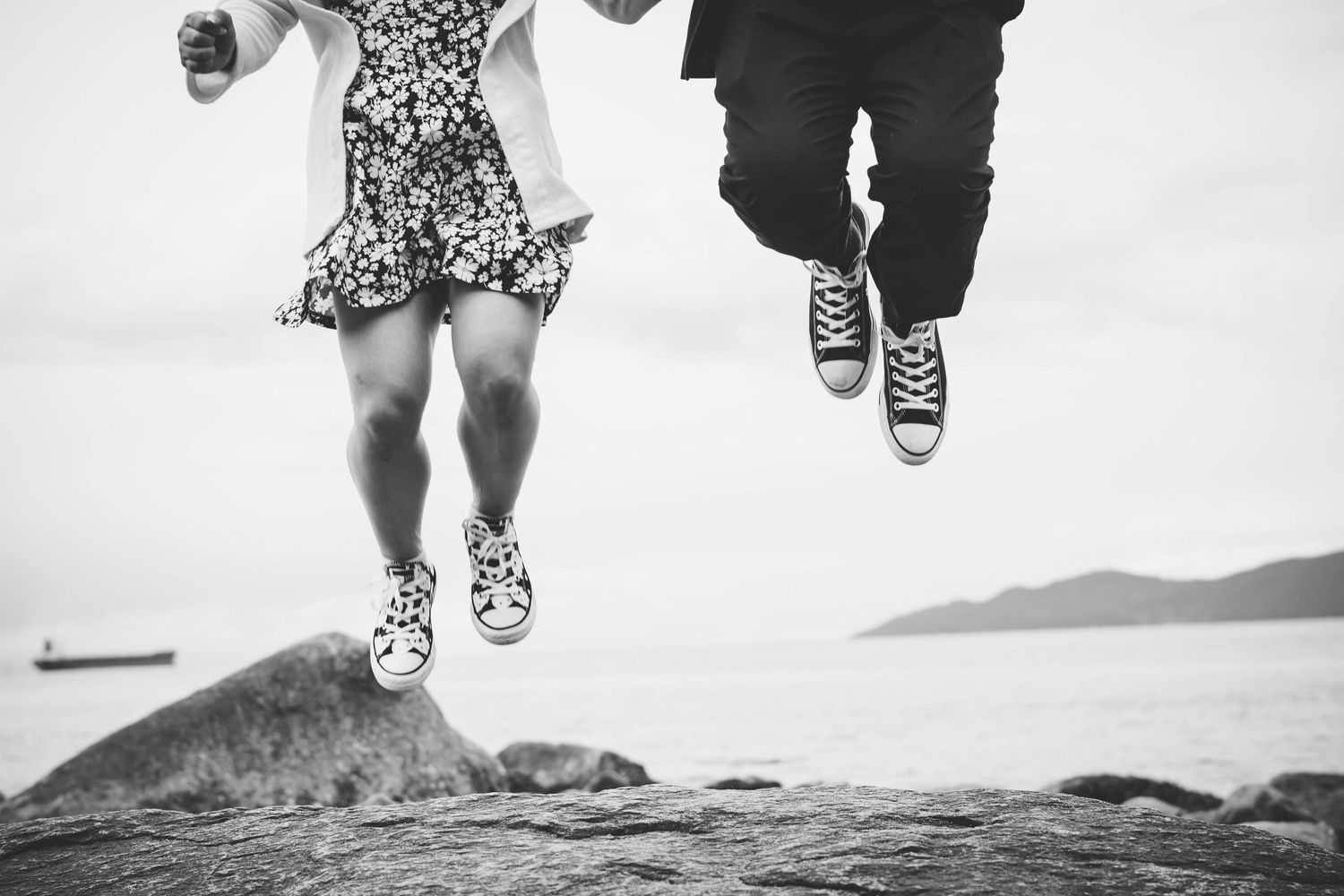 Black and white photo of two people jumping together on a cliff with mountains and water in the background.
