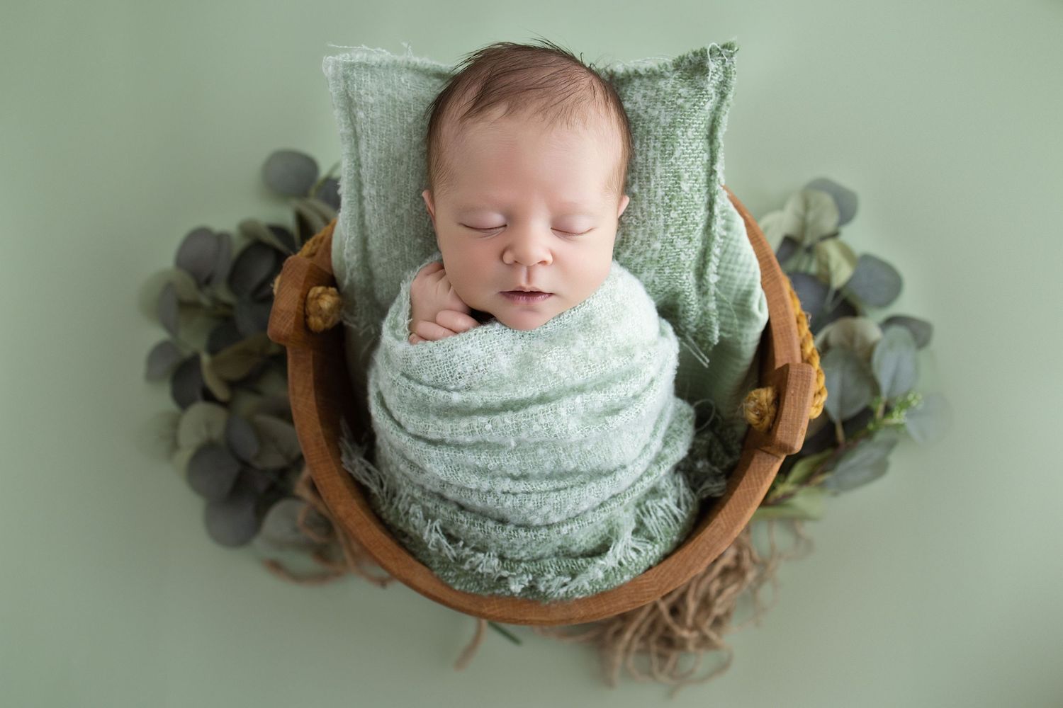 Newborn baby swaddled in mint green wrap sleeping peacefully in a wooden bowl with eucalyptus leaves.