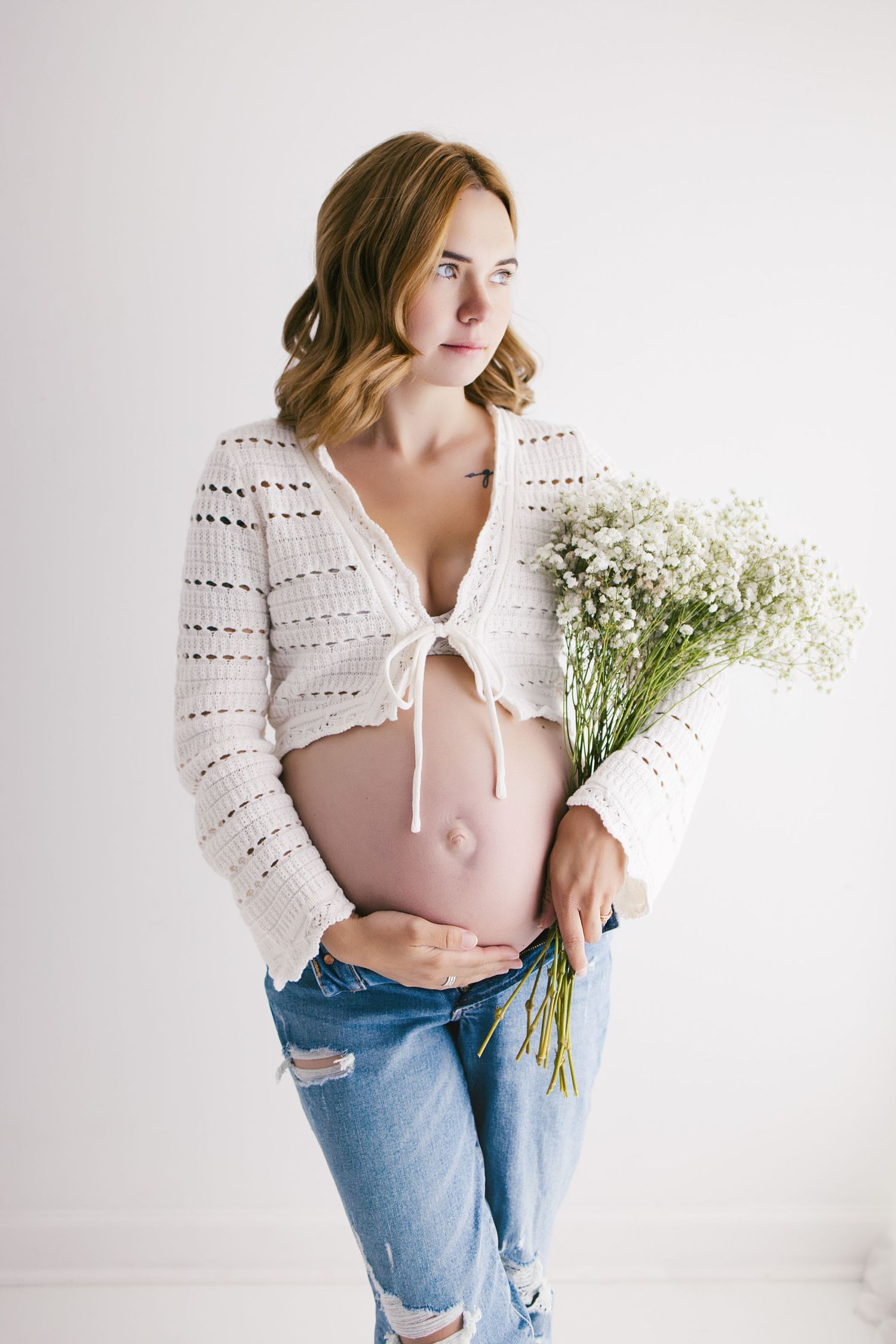 Pregnant woman in white crochet cardigan and ripped jeans holding white wildflowers, showcasing baby bump.
