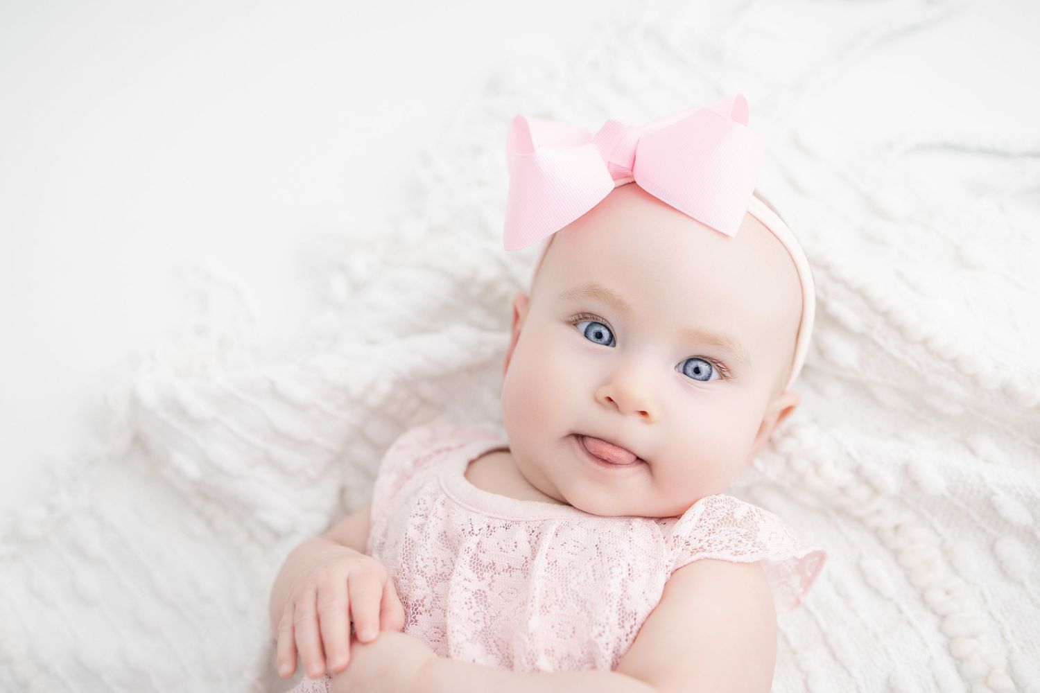 Adorable baby girl with blue eyes wearing a pink lace outfit and pink bow headband lying on a white blanket.