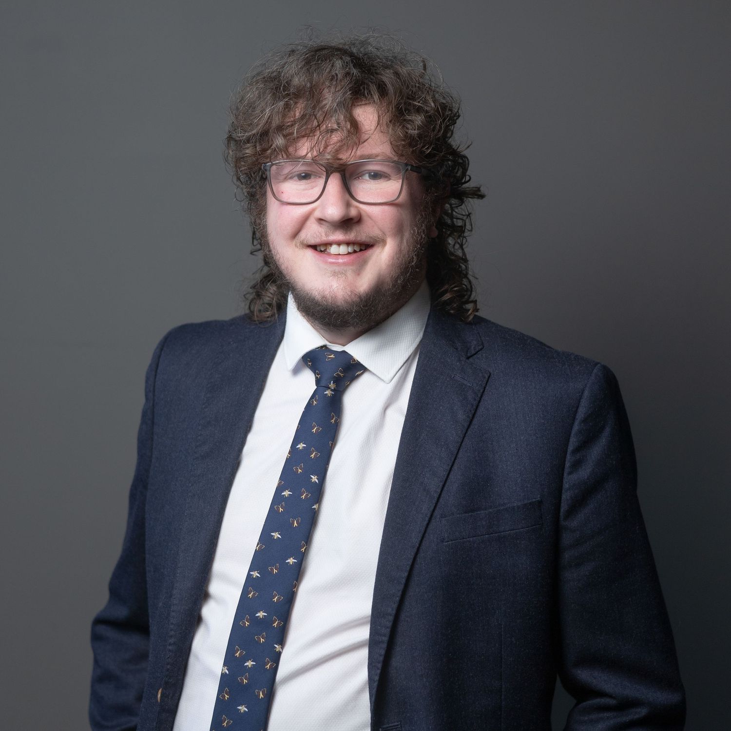 James in navy suit and patterned tie with curly hair and glasses, smiling against gray background. Aerial Site Intelligence
