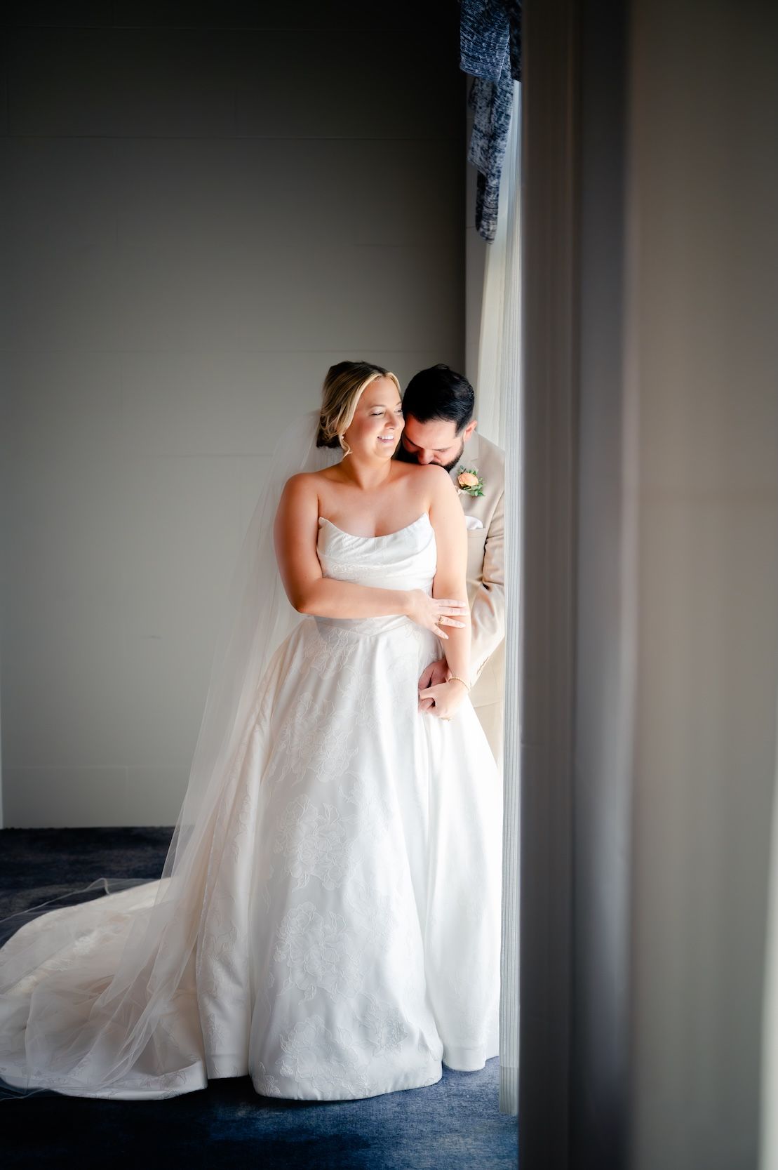 A bride in a white strapless gown embraced by a groom in a tan suit, standing by a window in soft natural light.