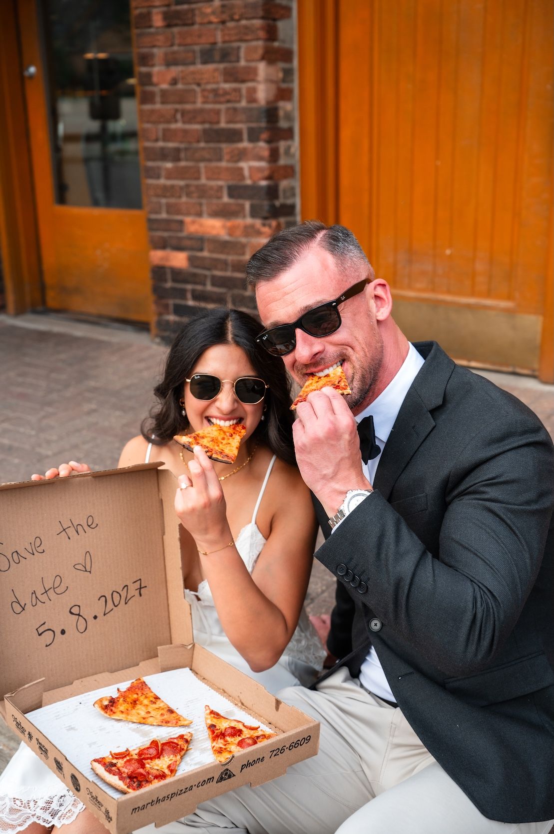 Couple in wedding attire eating pizza from a save the date box dated 5.8.2027, both wearing sunglasses.