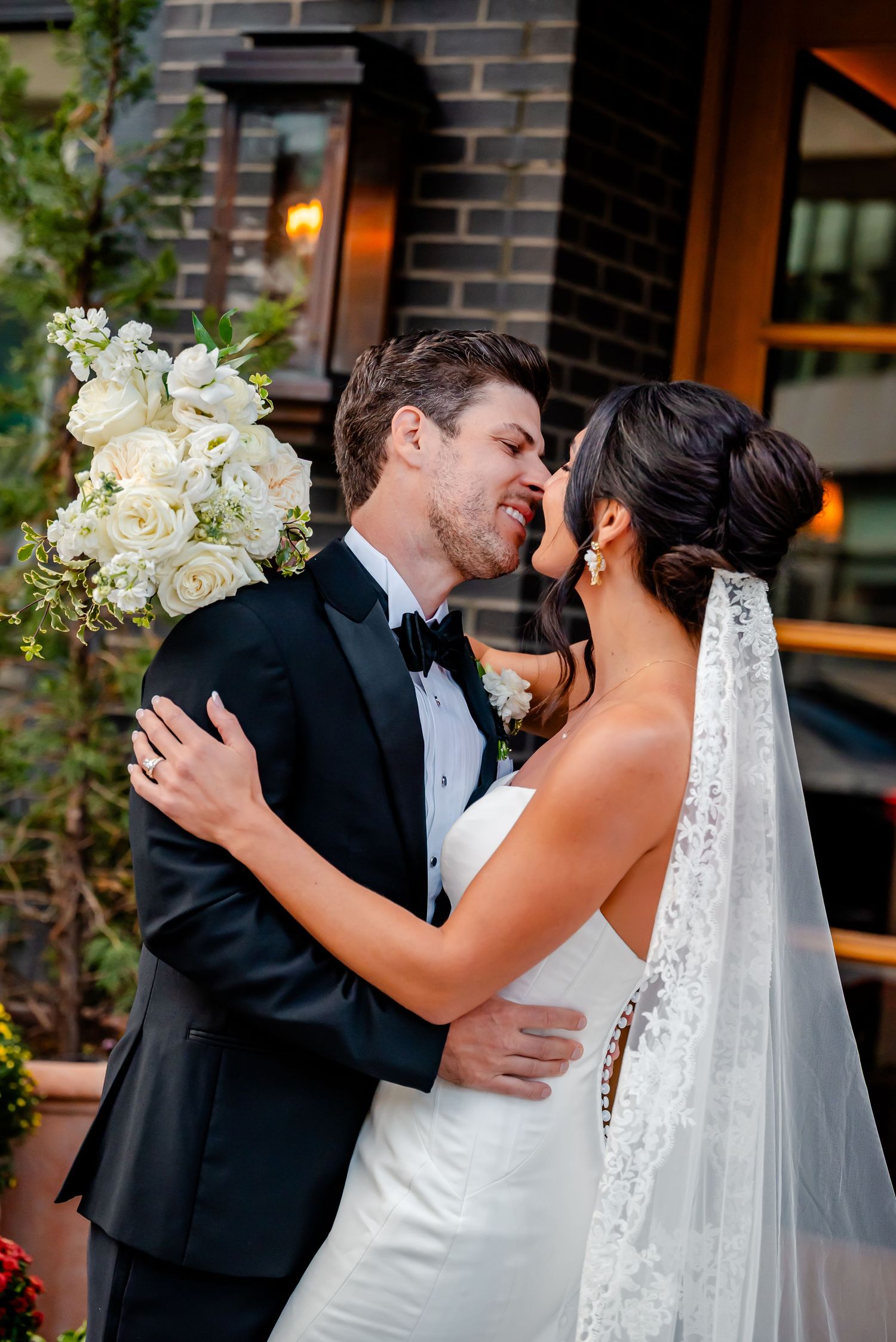 A bride and groom share a romantic moment in a close embrace, with white floral bouquet and lace veil outdoors.