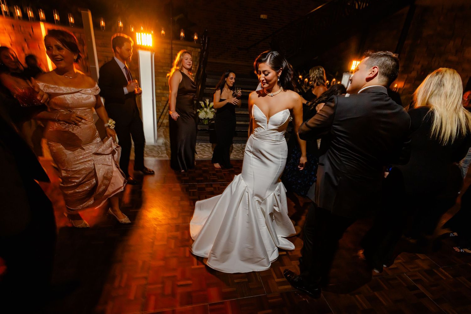 Bride in white mermaid gown dancing with guests at elegant wedding reception with warm ambient lighting.
