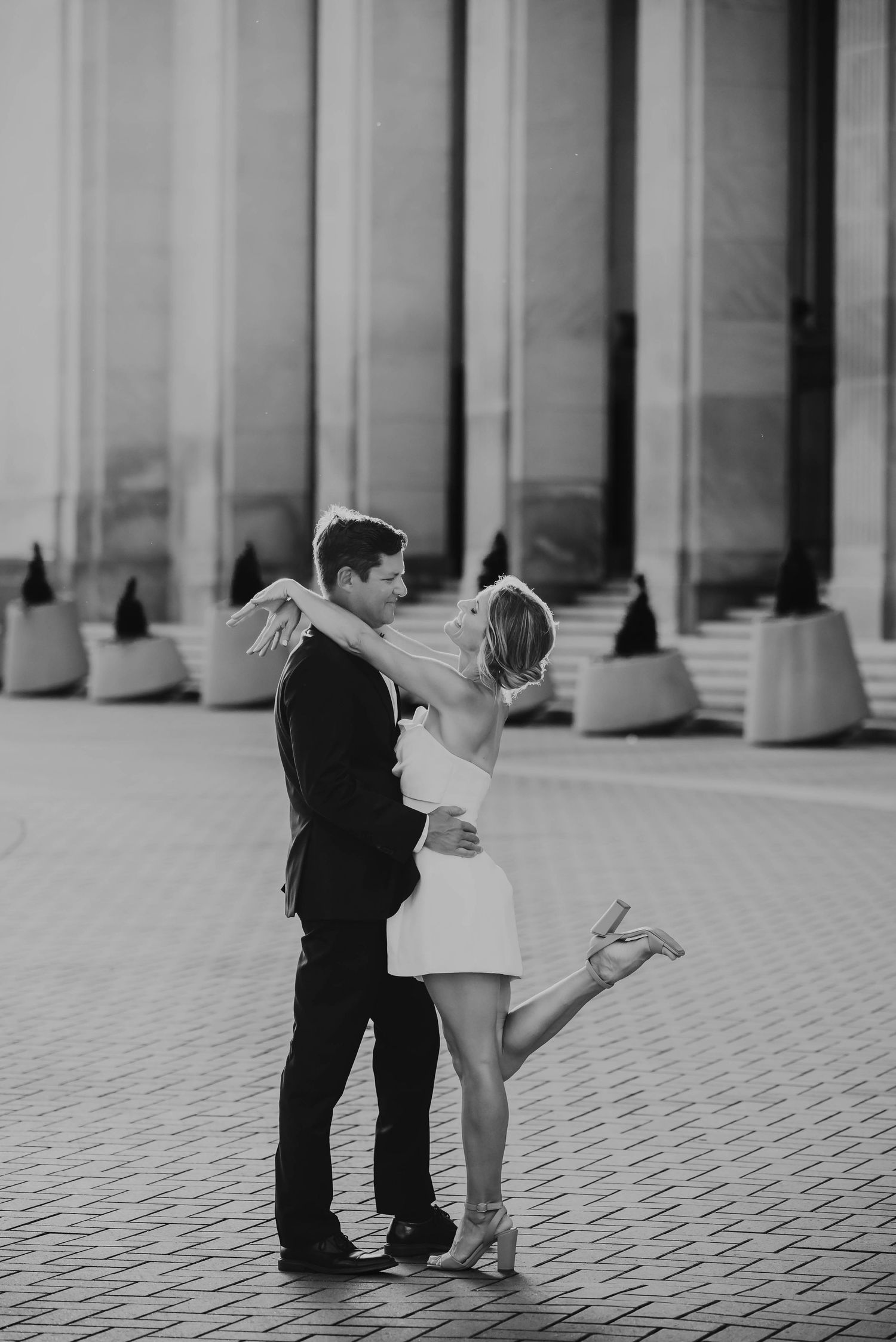 Black and white photo of a couple embracing and kissing outdoors near a modern building with tall columns at The Amway Hotel in Grand Rapids