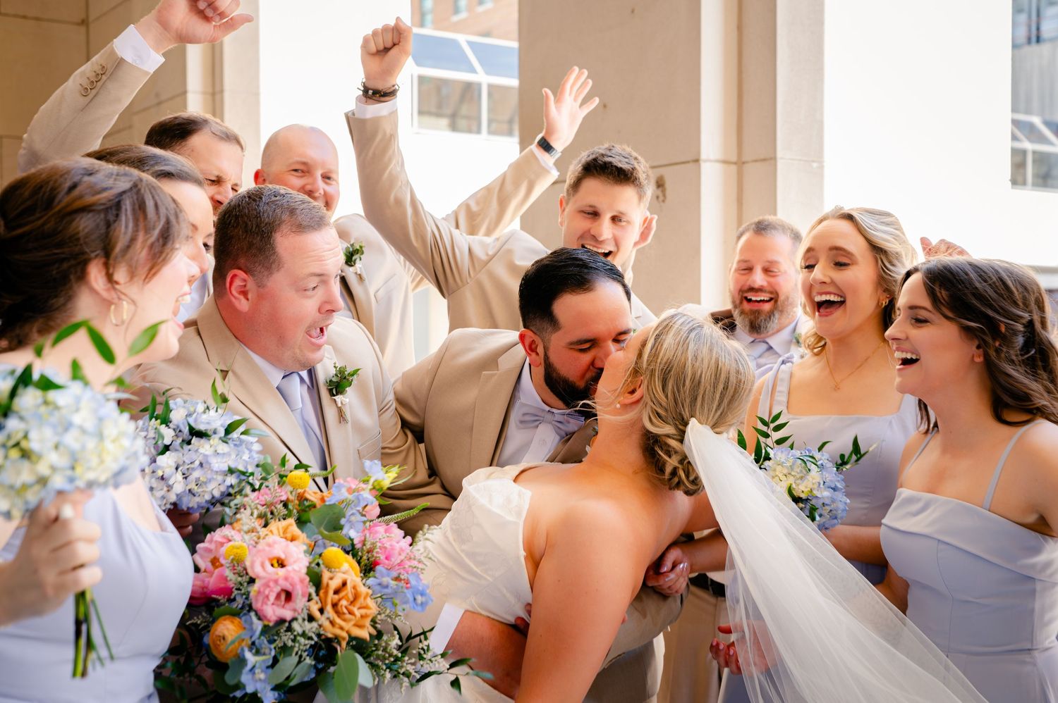 Wedding party cheering as bride and groom share a kiss, surrounded by bridesmaids and groomsmen celebrating outdoors.
