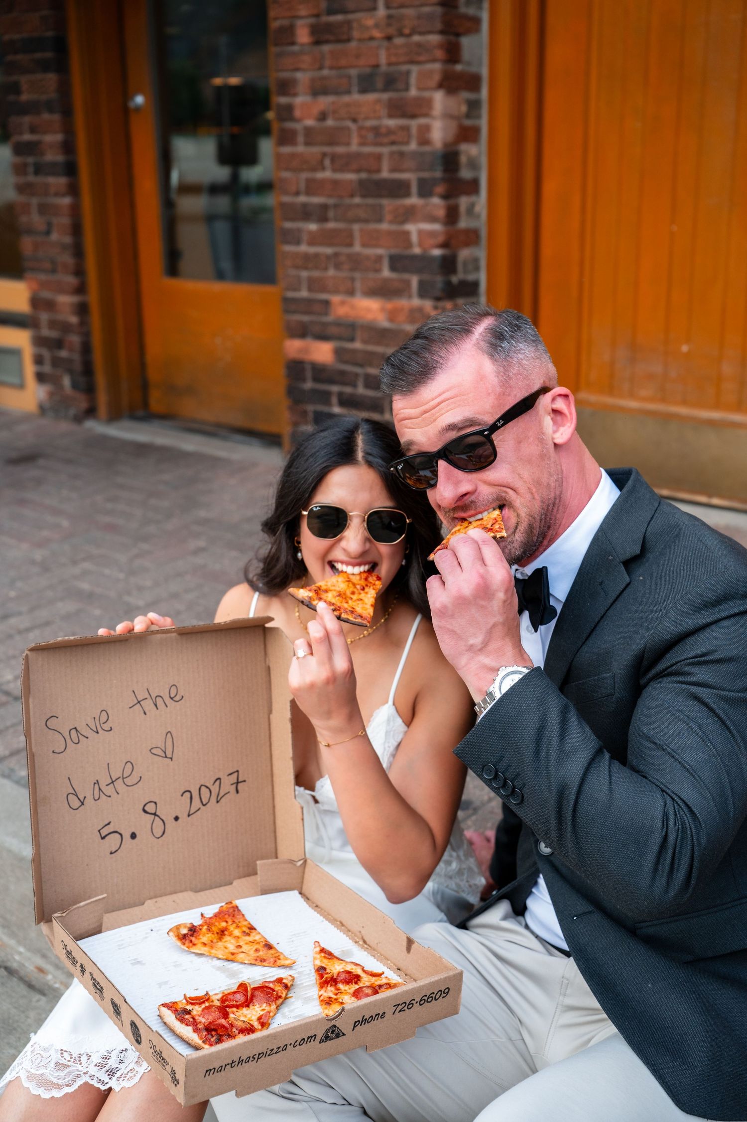 Couple in wedding attire eating pizza from a box with Save the Date 5.8.2027 written on it.