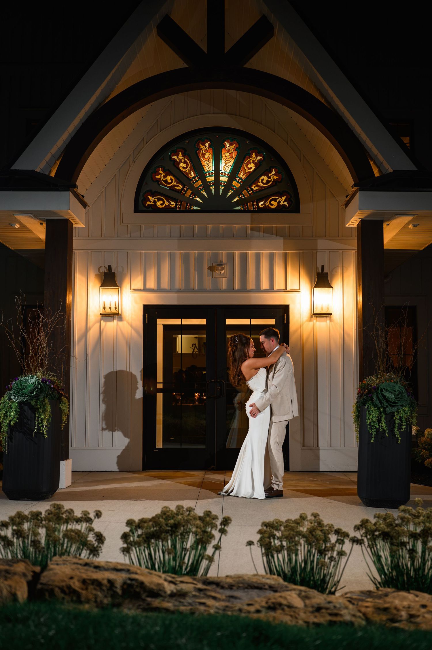 Couple sharing a romantic kiss at night outside an elegant white chapel entrance with stained glass and warm lighting.