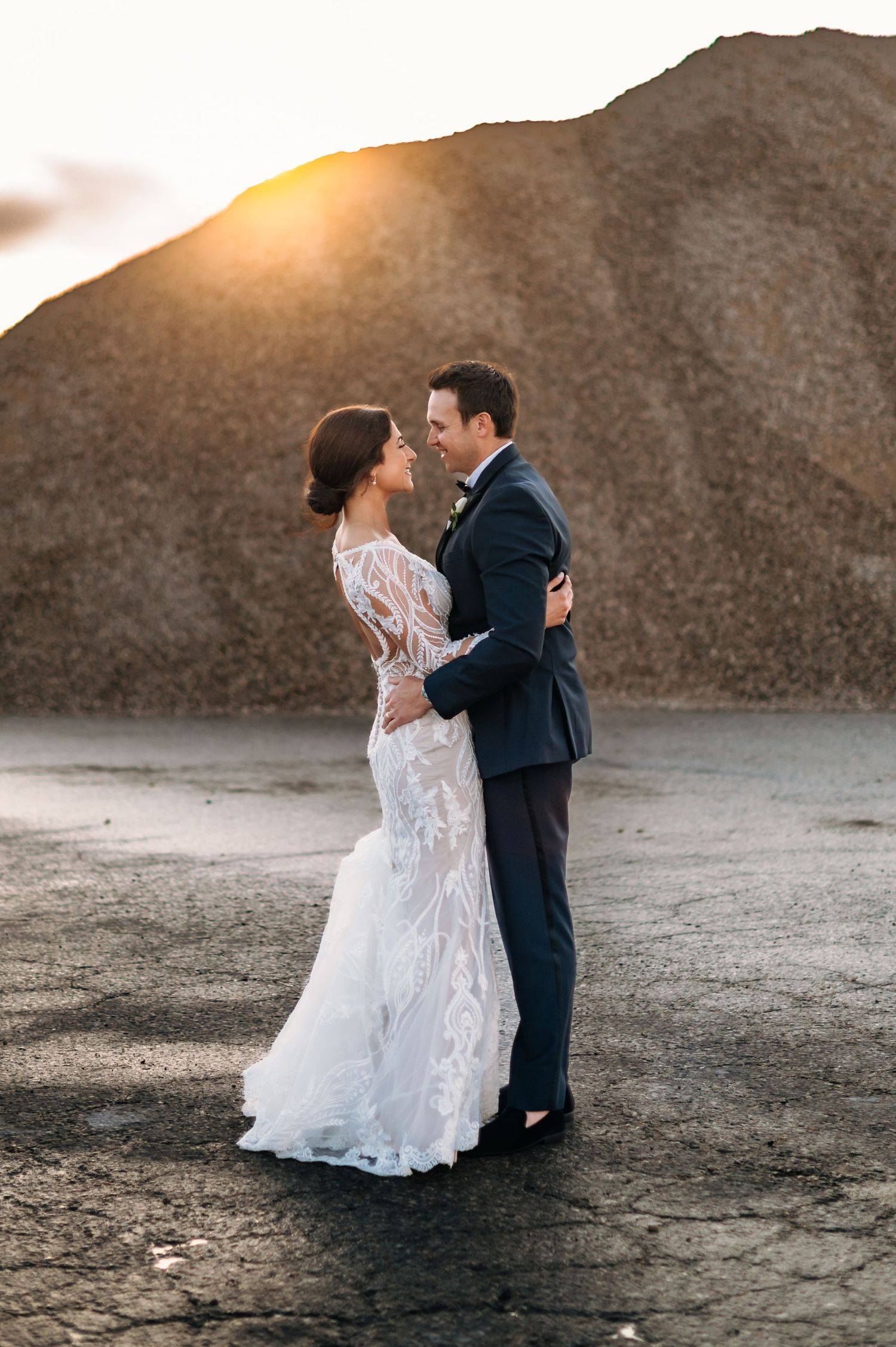 Romantic wedding couple embraces at sunset against rugged mountainous backdrop, bride in lace gown and groom in navy suit.