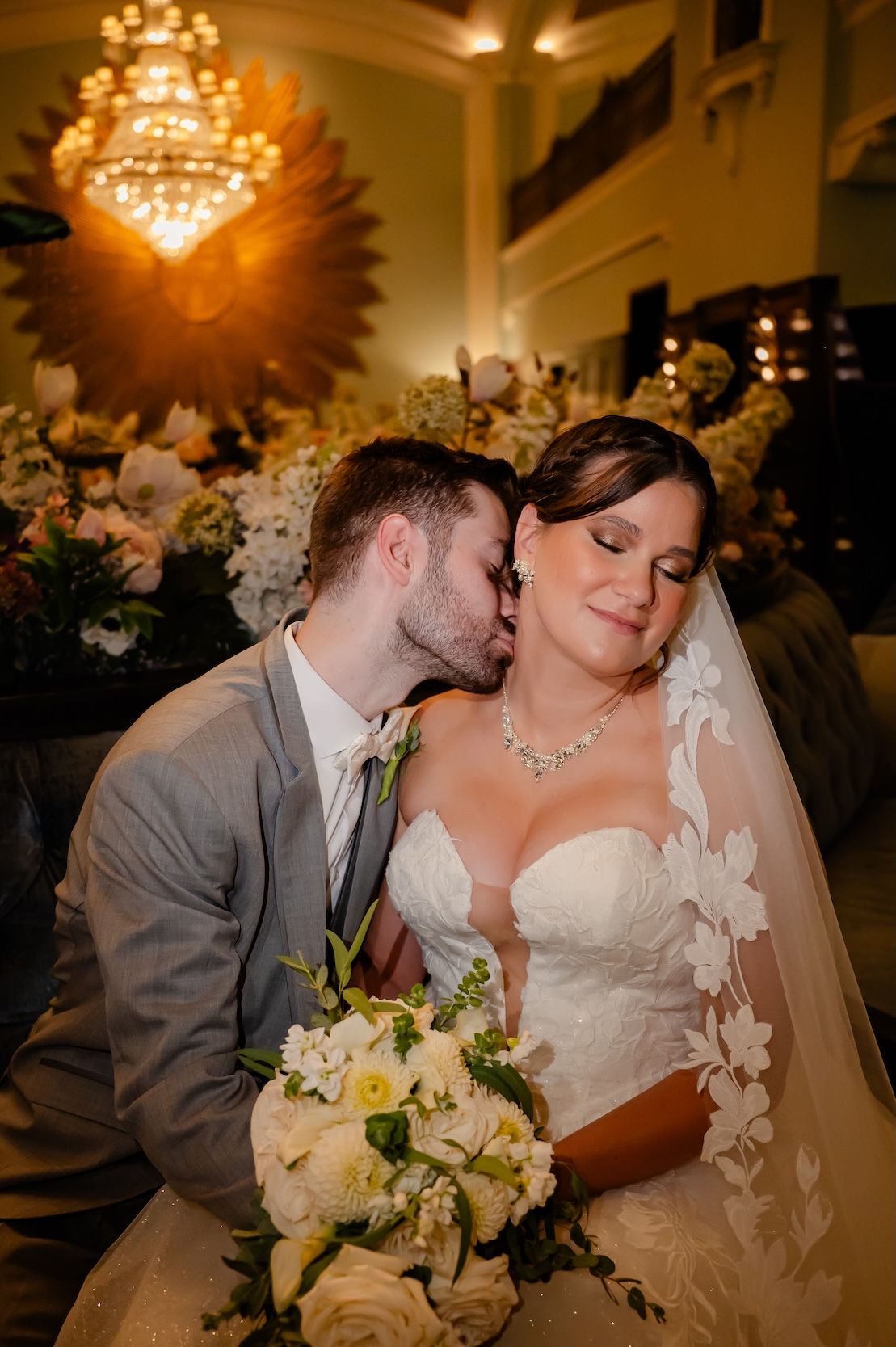 Groom in gray suit kisses bride in lace wedding dress holding white bouquet under chandelier.