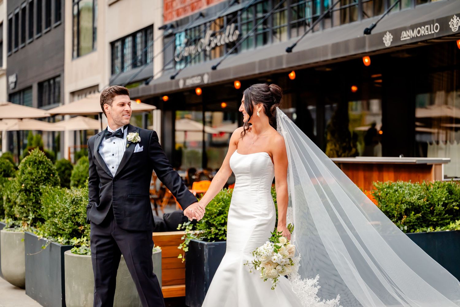 Bride and groom hold hands outdoors near urban restaurant, bride wearing strapless gown with long veil, groom in black tuxedo.