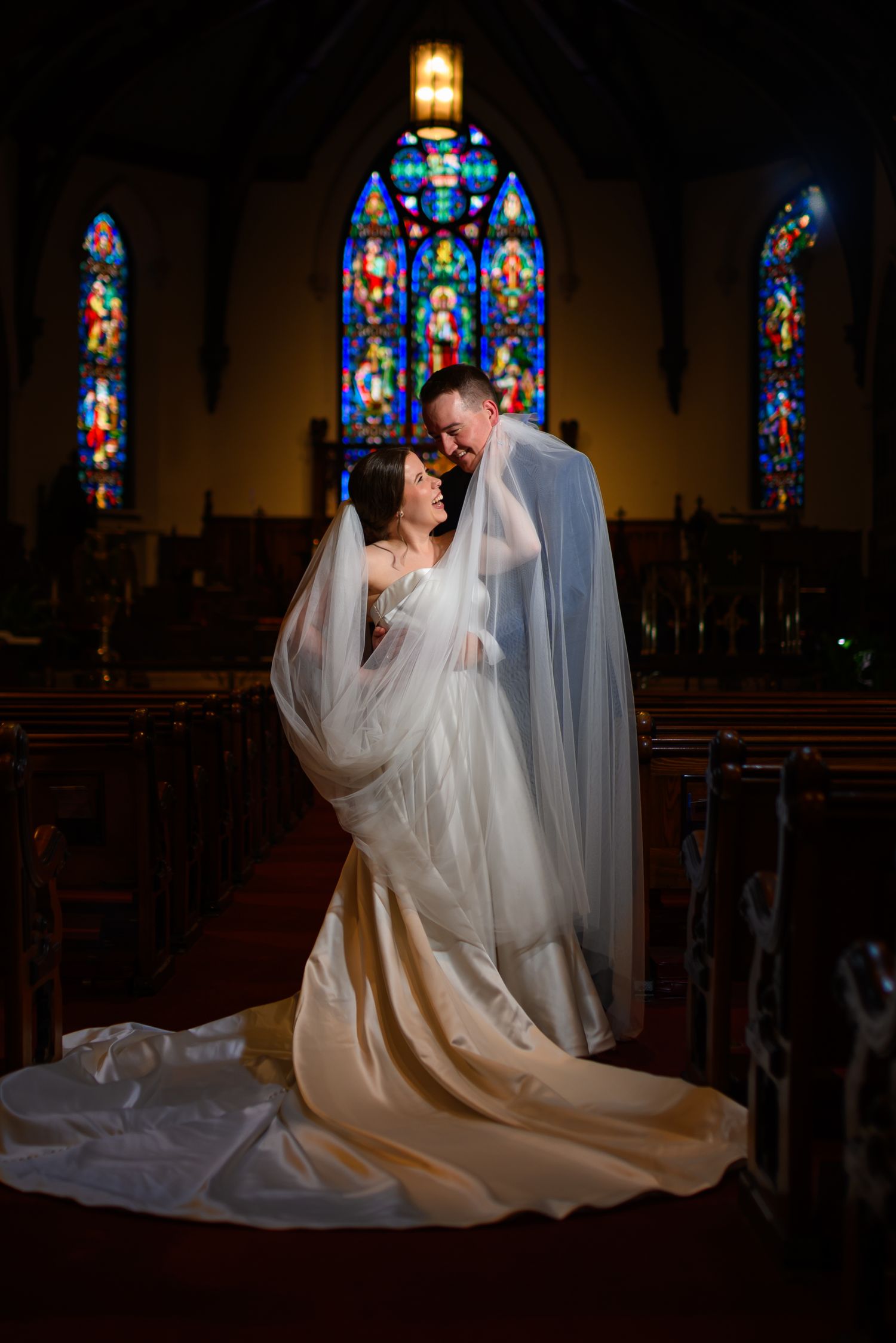 A wedding couple embraces in a dramatic pose inside a church with beautiful stained glass windows in the background.