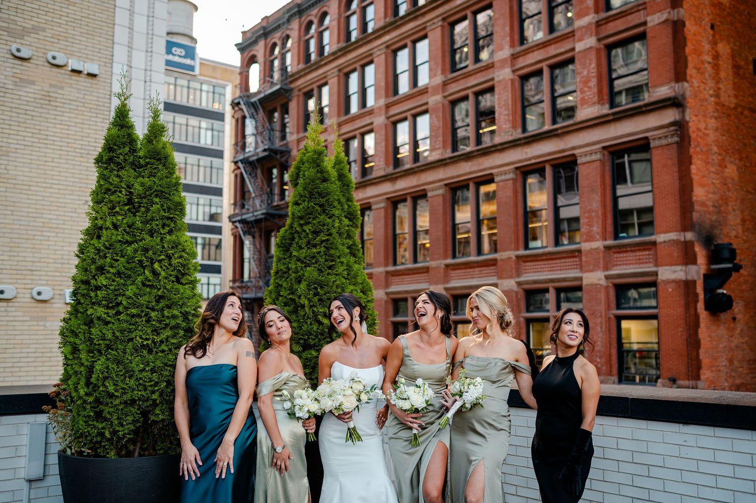 Bride with bridesmaids in sage and navy dresses laughing on rooftop terrace with brick building backdrop.