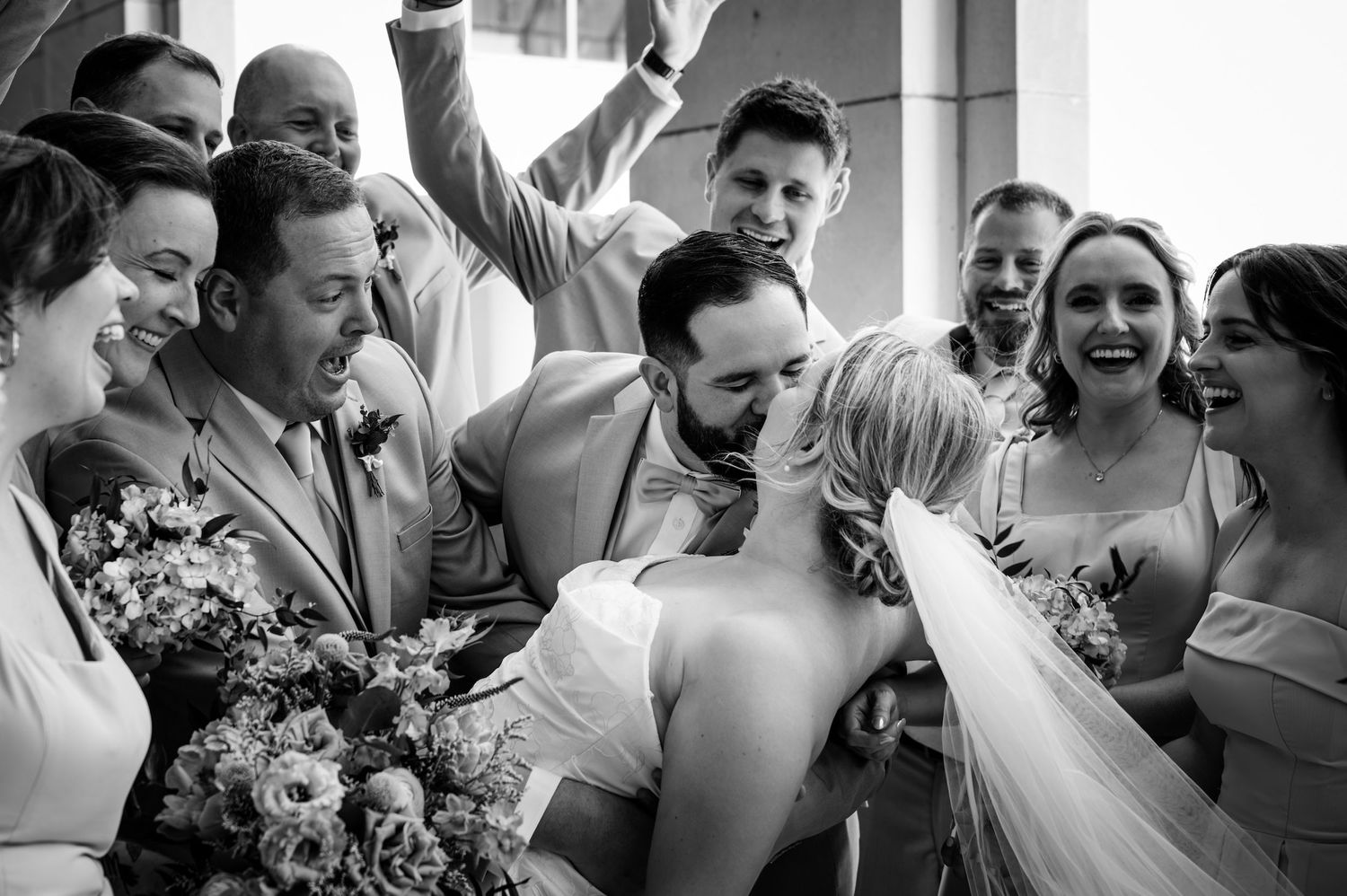 Black and white photo of bride and groom kissing surrounded by cheering wedding party guests.