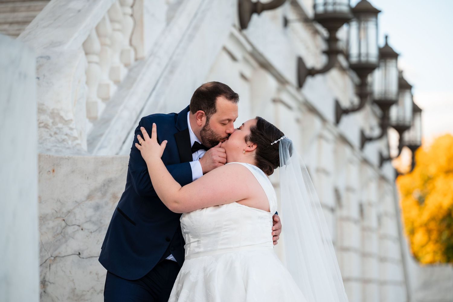Bride and groom sharing a romantic kiss outdoors near elegant white stone columns on their wedding day.