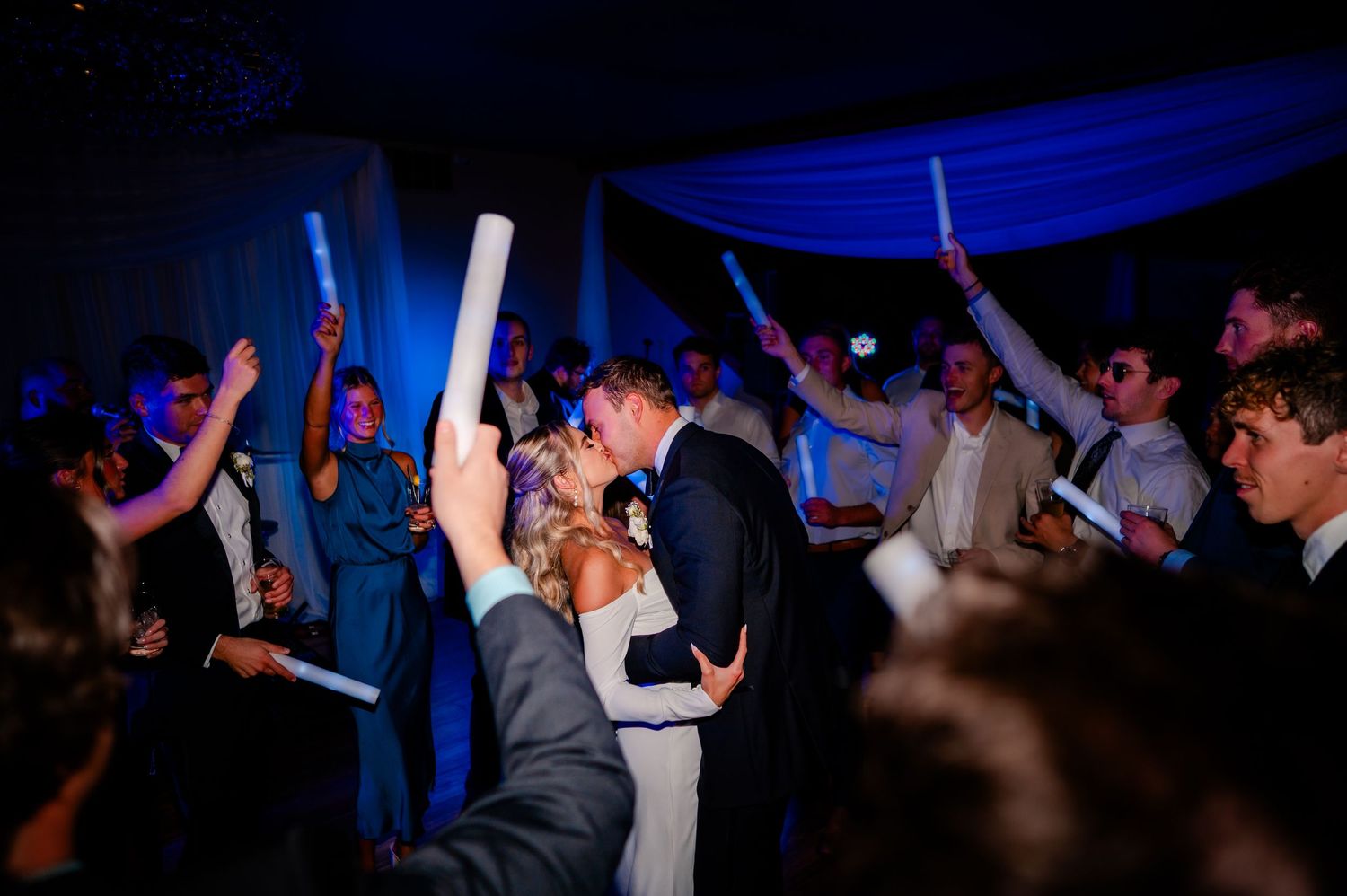 Wedding guests dance with glowing blue LED foam sticks on a dark dance floor at a reception celebration.