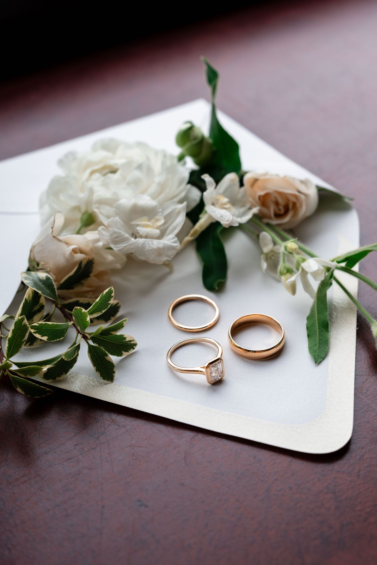 Three gold wedding rings beside white and blush flowers on an envelope, including a solitaire diamond engagement ring.