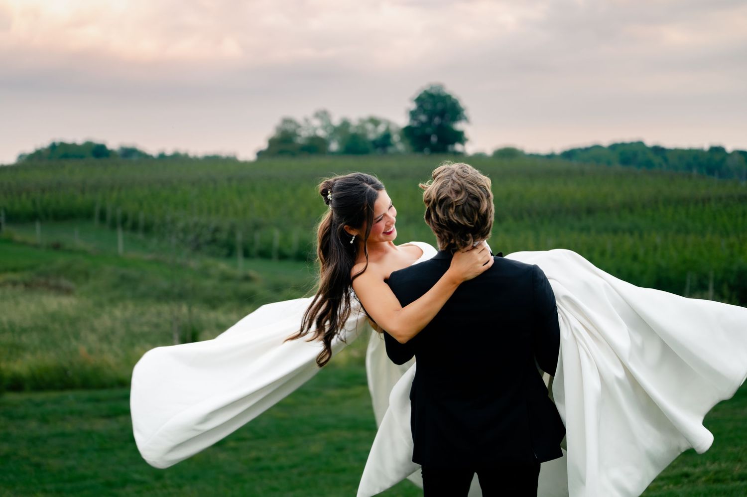A bride in a flowing white gown and groom in black suit share a romantic moment at a vineyard wedding at sunset.