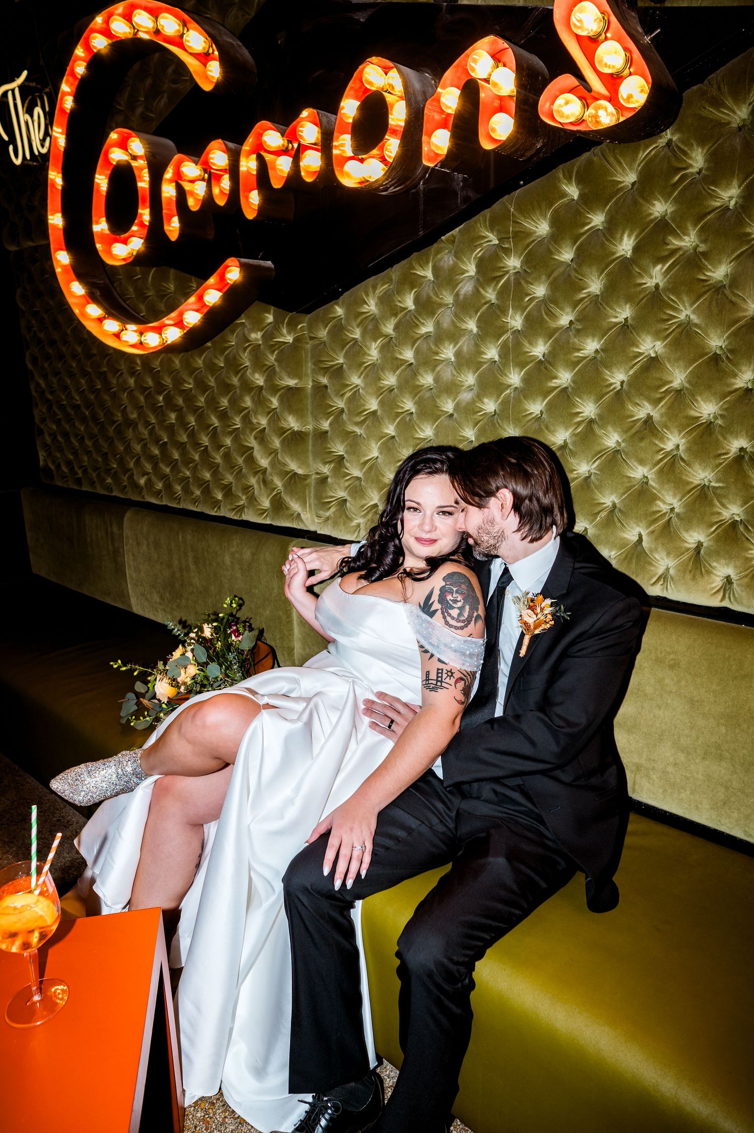 A tattooed bride in white sits on her groom's lap at The Commons venue, sharing a romantic moment in a tufted gold booth.