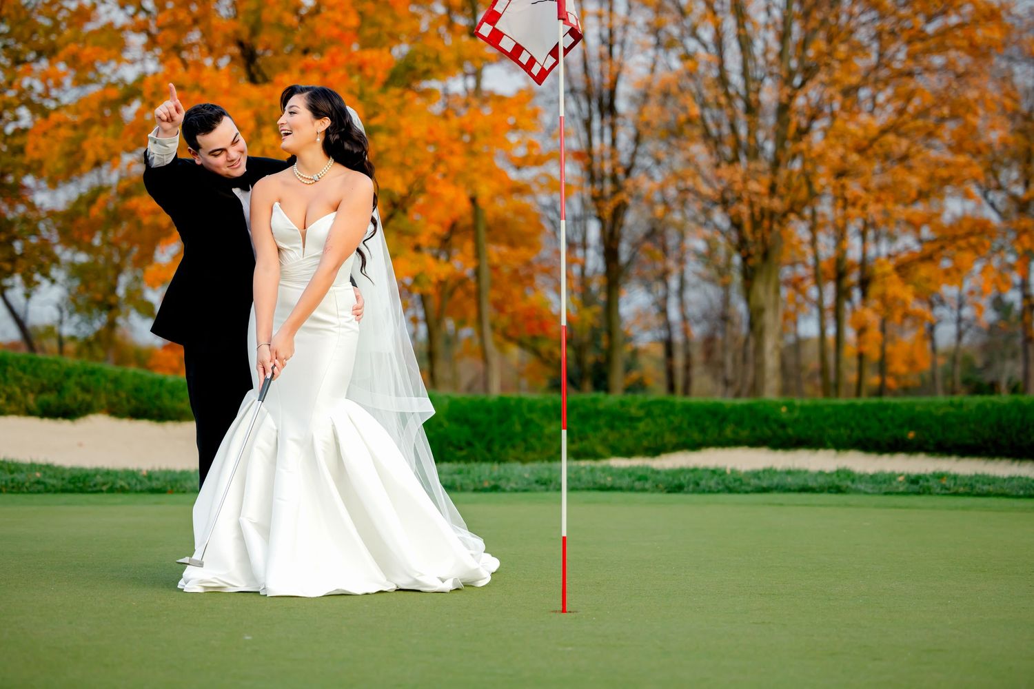 Bride and groom pose on a golf course green with autumn foliage backdrop, groom helping bride with a golf swing.