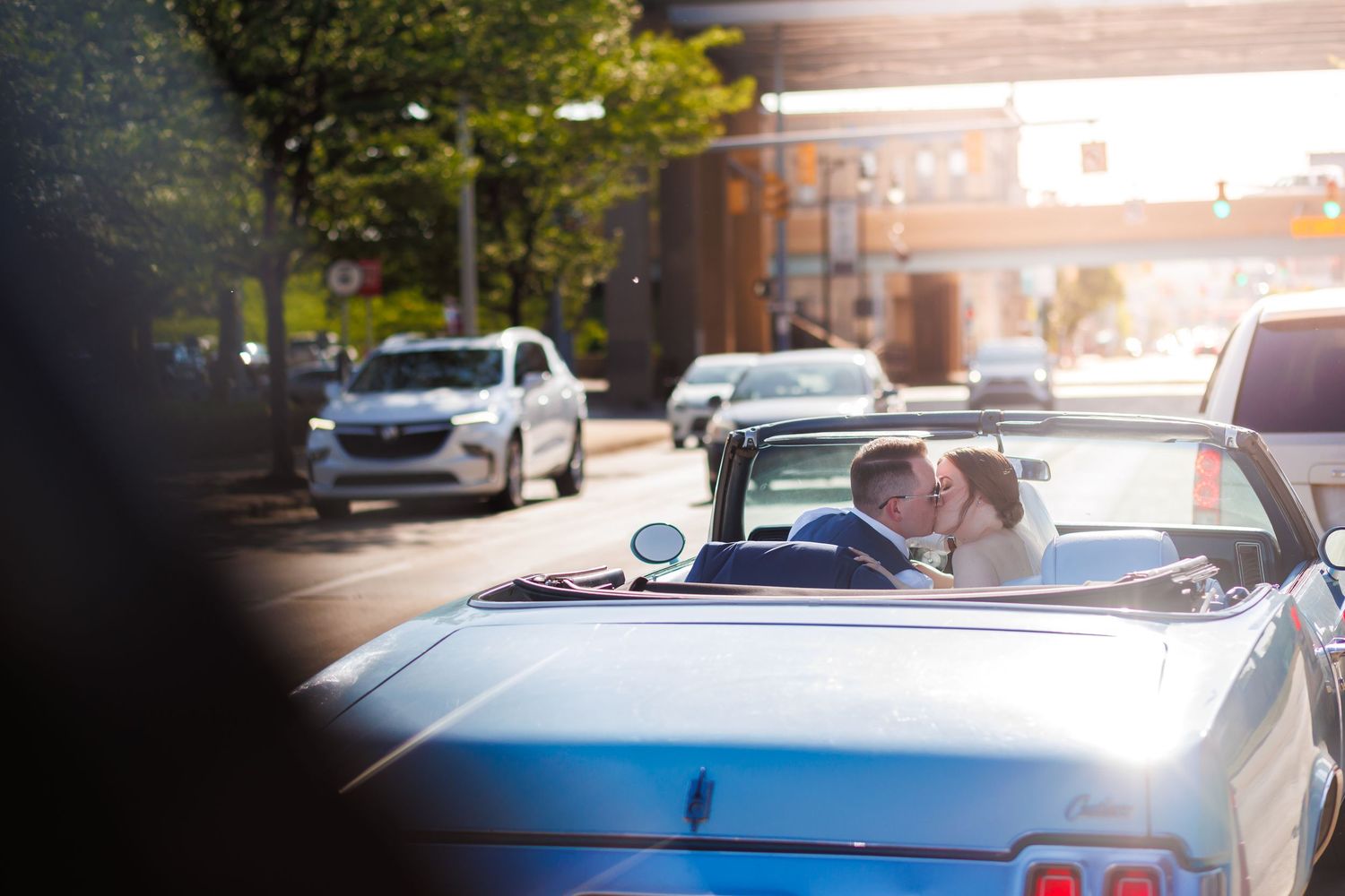 Newlywed couple sharing a kiss in a classic convertible car on a sunny city street.