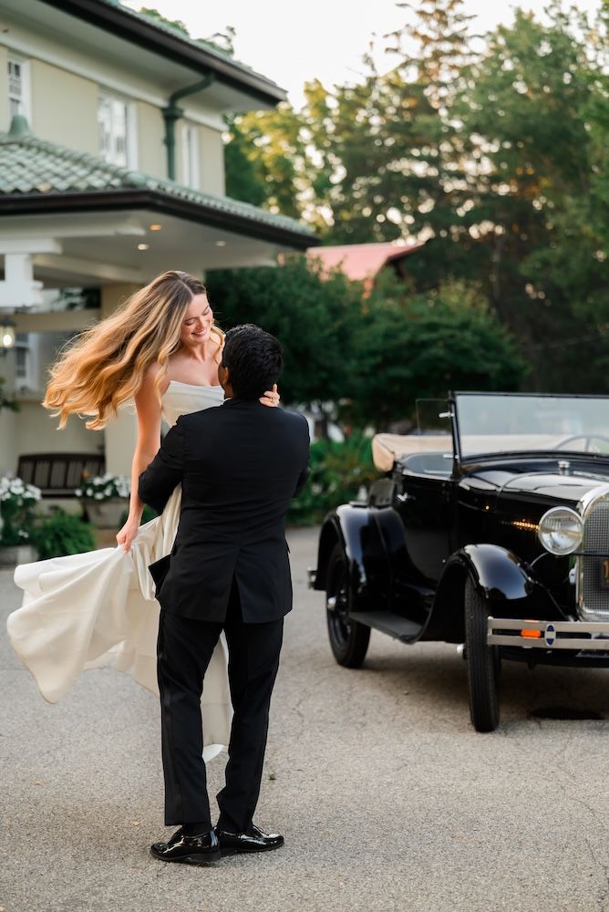 Groom lifting bride in white dress near vintage black car outside elegant venue at golden hour.