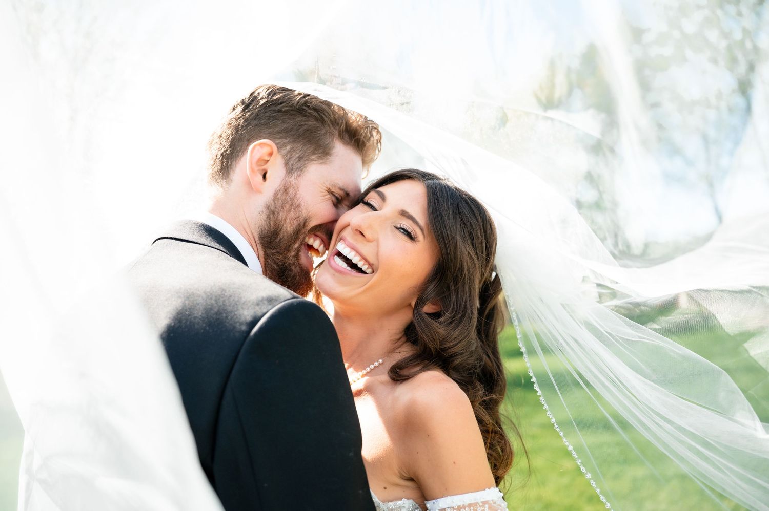 Joyful bride and groom laughing under flowing white veil on sunny wedding day.