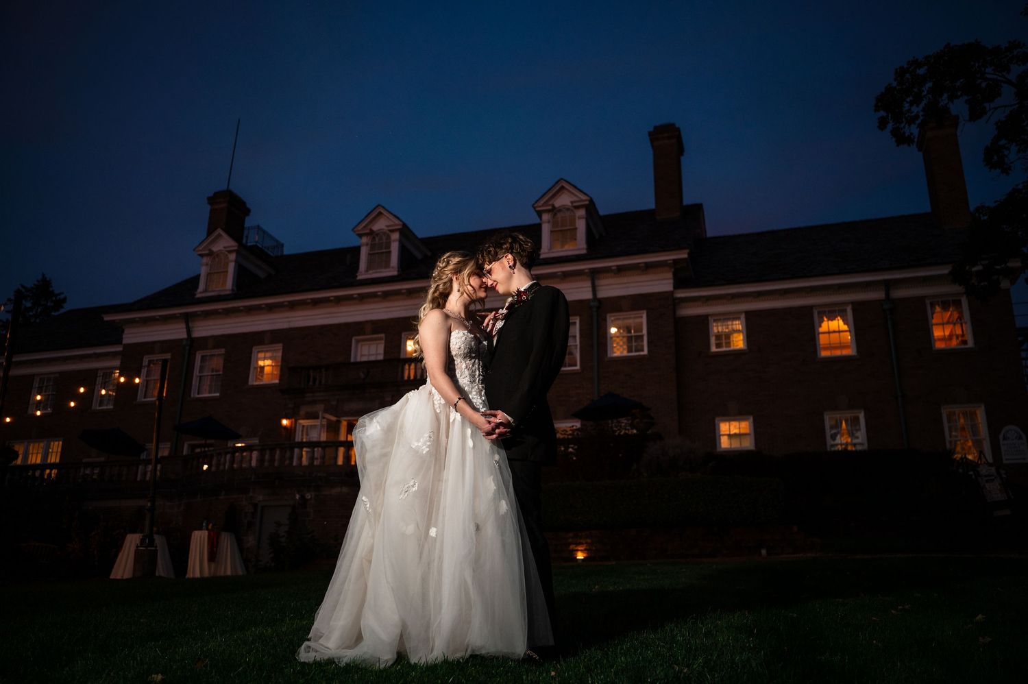 Bride and groom share a romantic kiss at dusk outside an elegant lit venue during their evening wedding celebration.