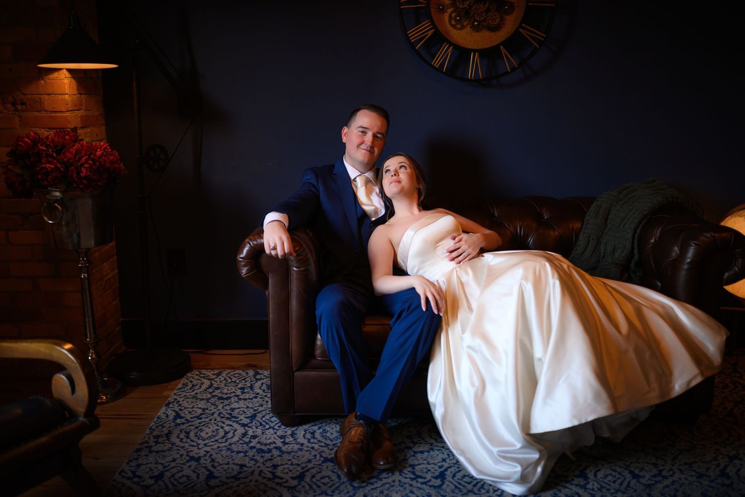 Bride in white gown reclines on groom in blue suit on leather sofa in elegant dark blue room.