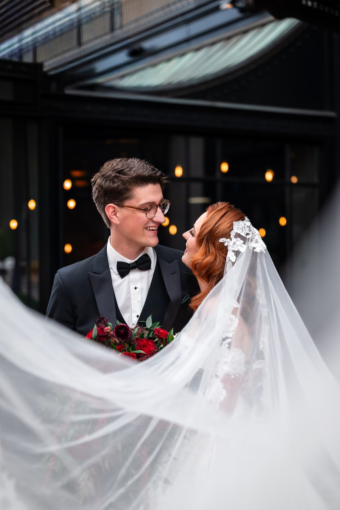 A smiling groom in a tuxedo gazes at his red-haired bride in a lace veil holding red roses outdoors.
