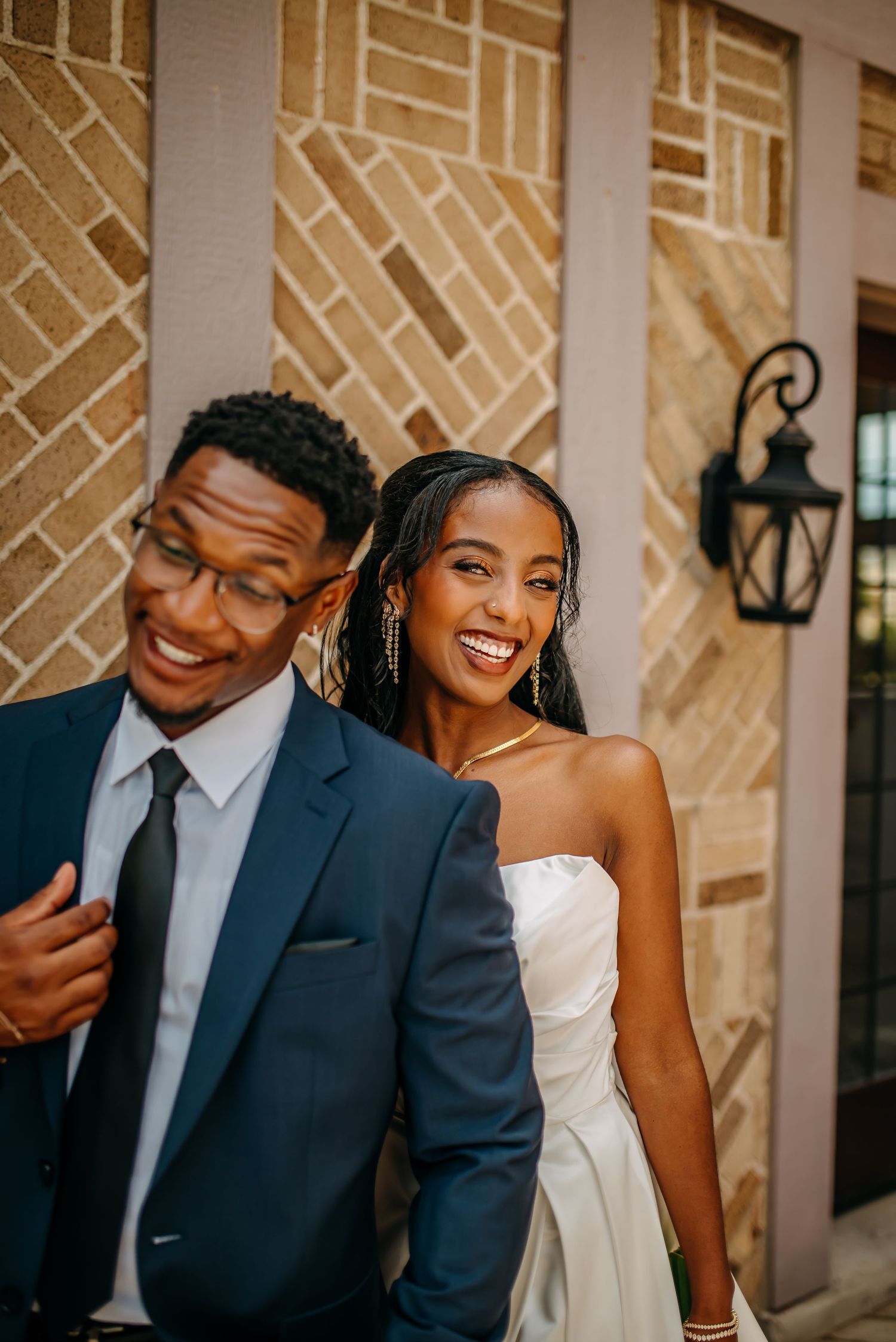 Smiling bride and groom in wedding attire pose together against a brick wall backdrop.