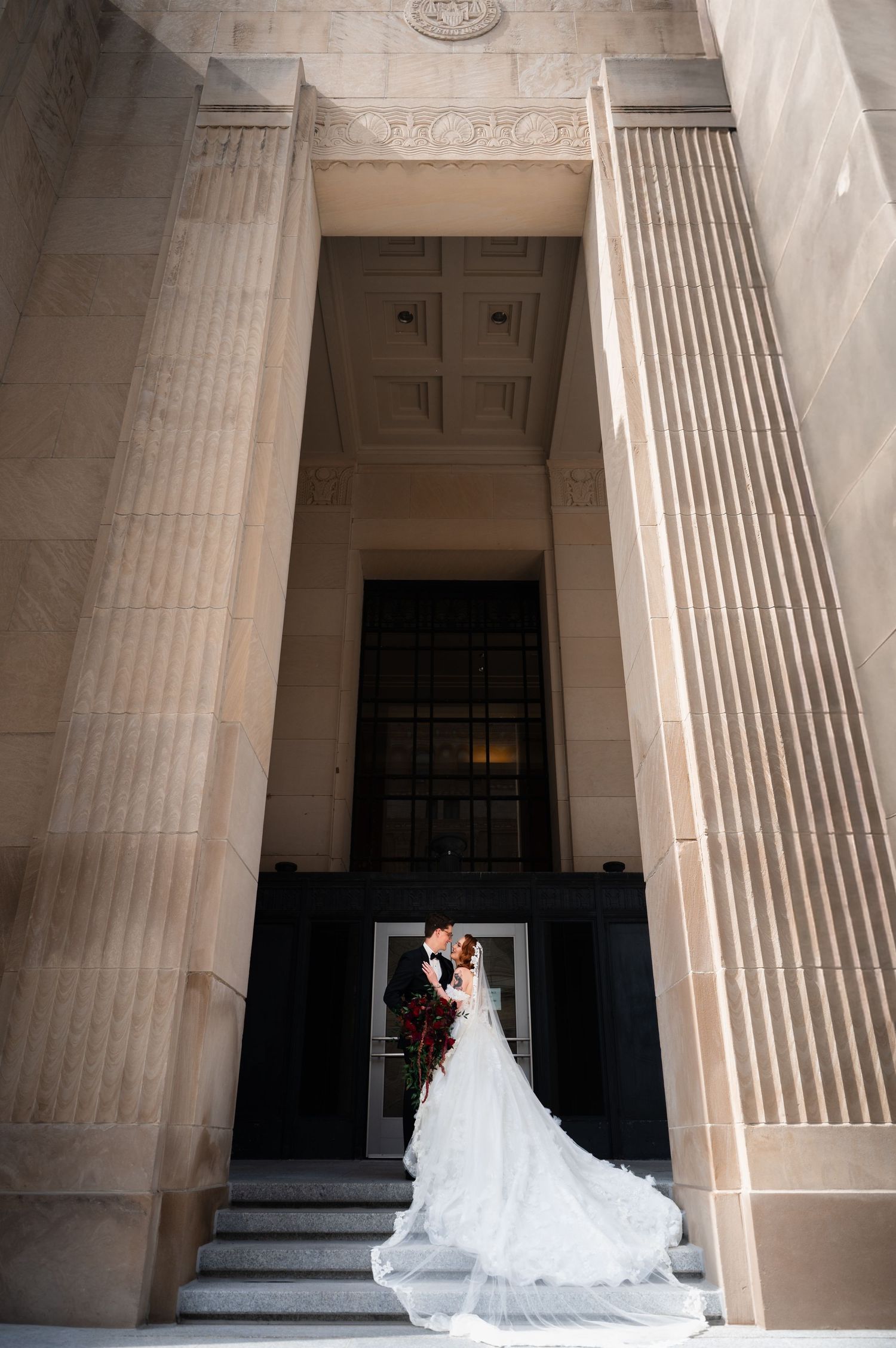 Bride and groom embrace on steps of grand neoclassical building with tall columns and flowing white wedding gown train.