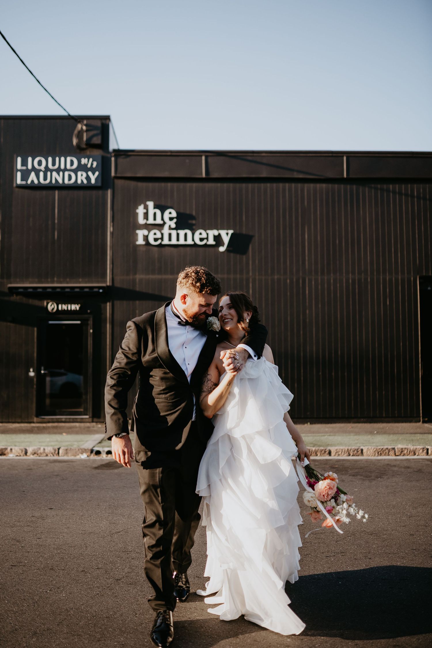 A couple in wedding attire share a romantic moment outside The Refinery and Liquid Laundry buildings.