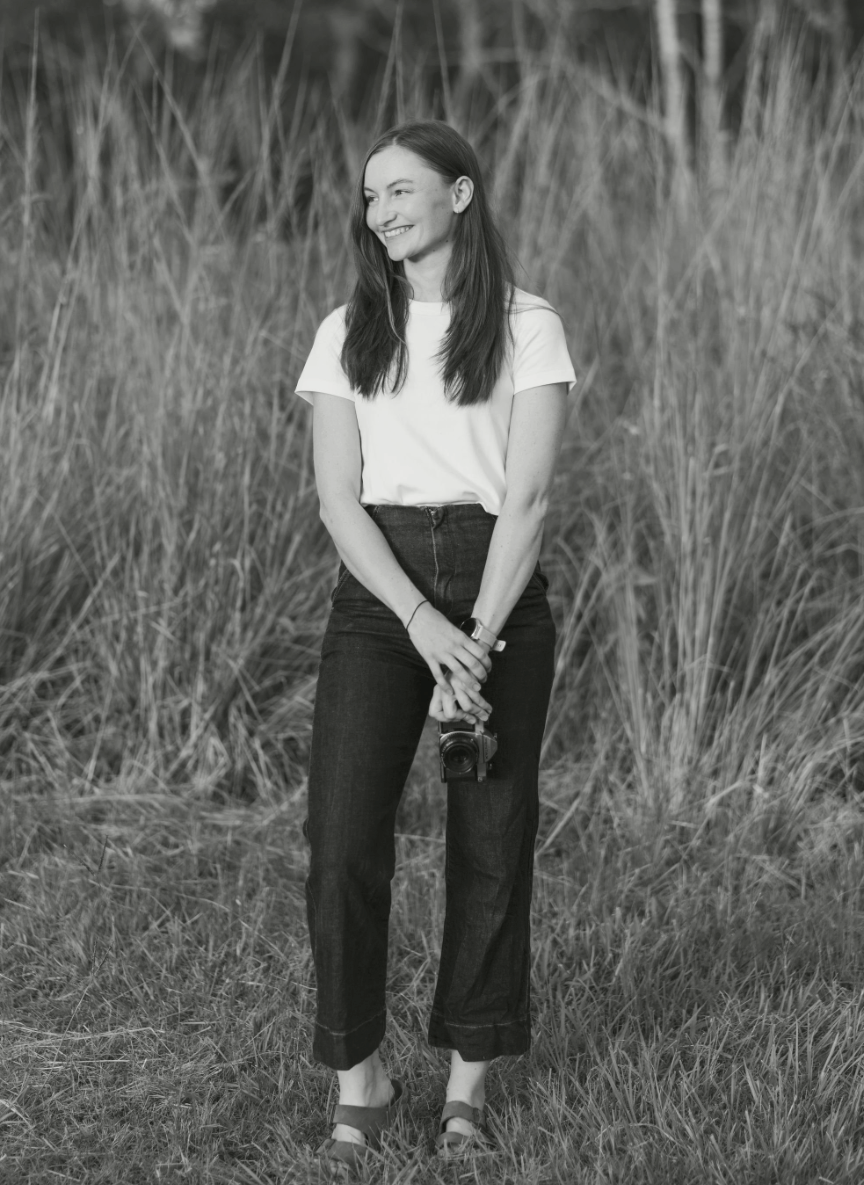 Black and white portrait of someone in a white t-shirt and dark pants standing in tall grass with a natural smile.