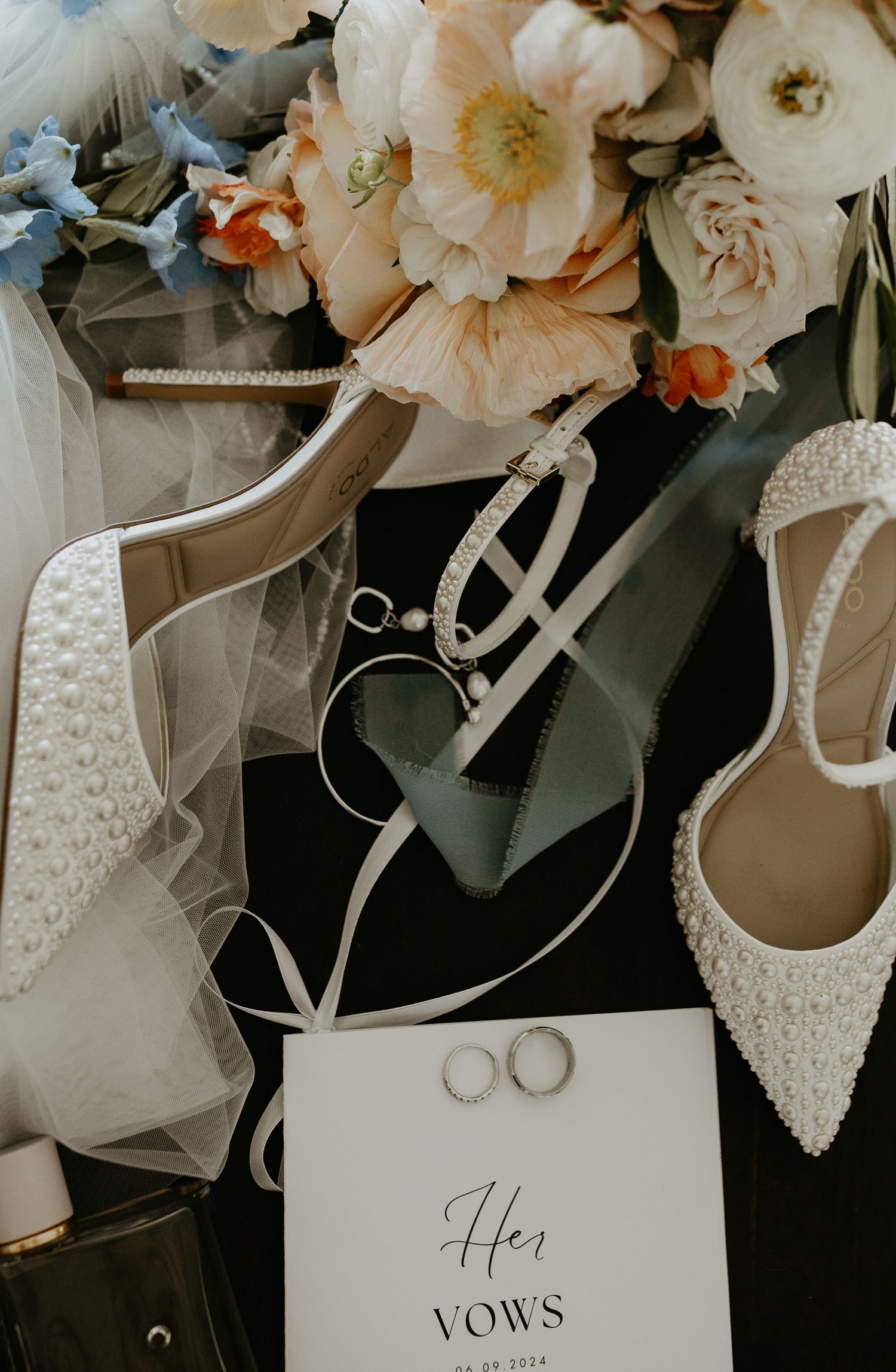Wedding details flatlay with white bridal shoes, floral bouquet, wedding bands and vows card in soft muted tones.