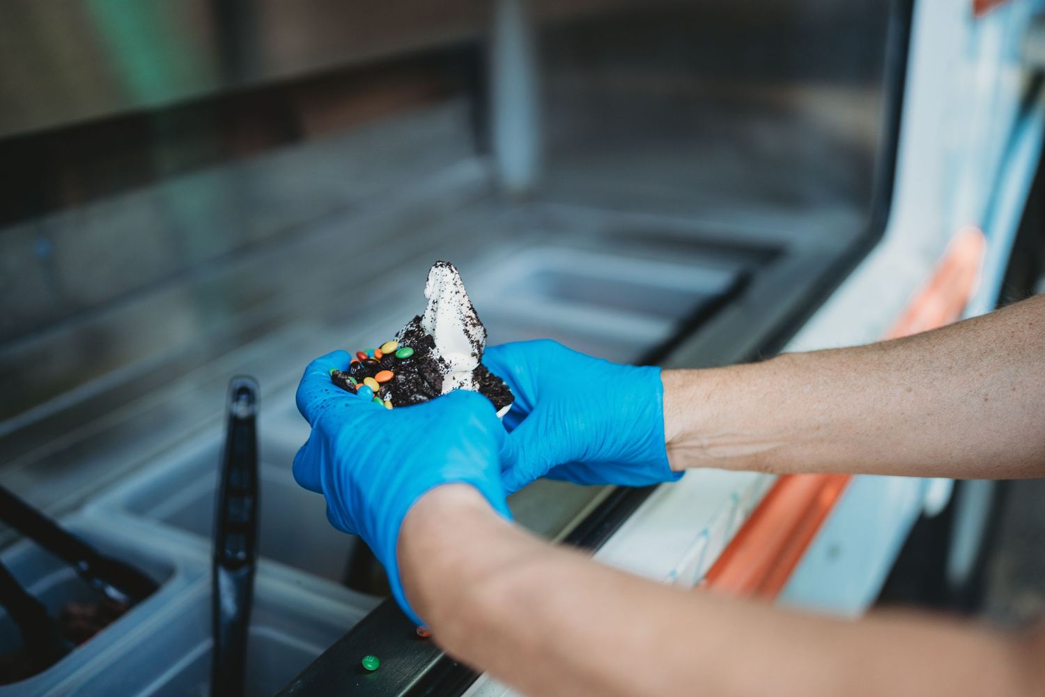 Preparing ice cream dessert with toppings while wearing protective blue gloves.