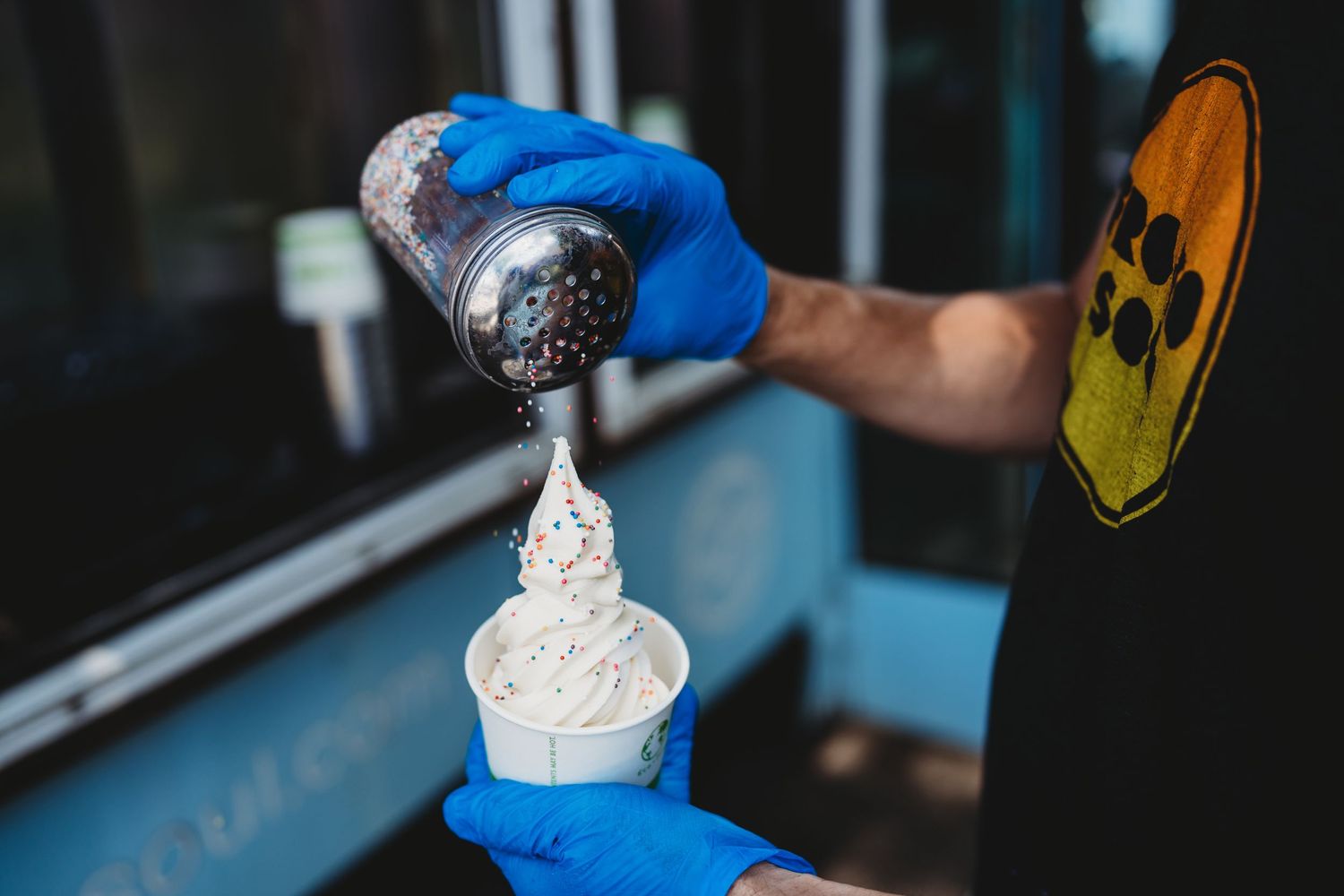 Ice cream being dispensed into a paper cup with blue gloved hands holding toppings.
