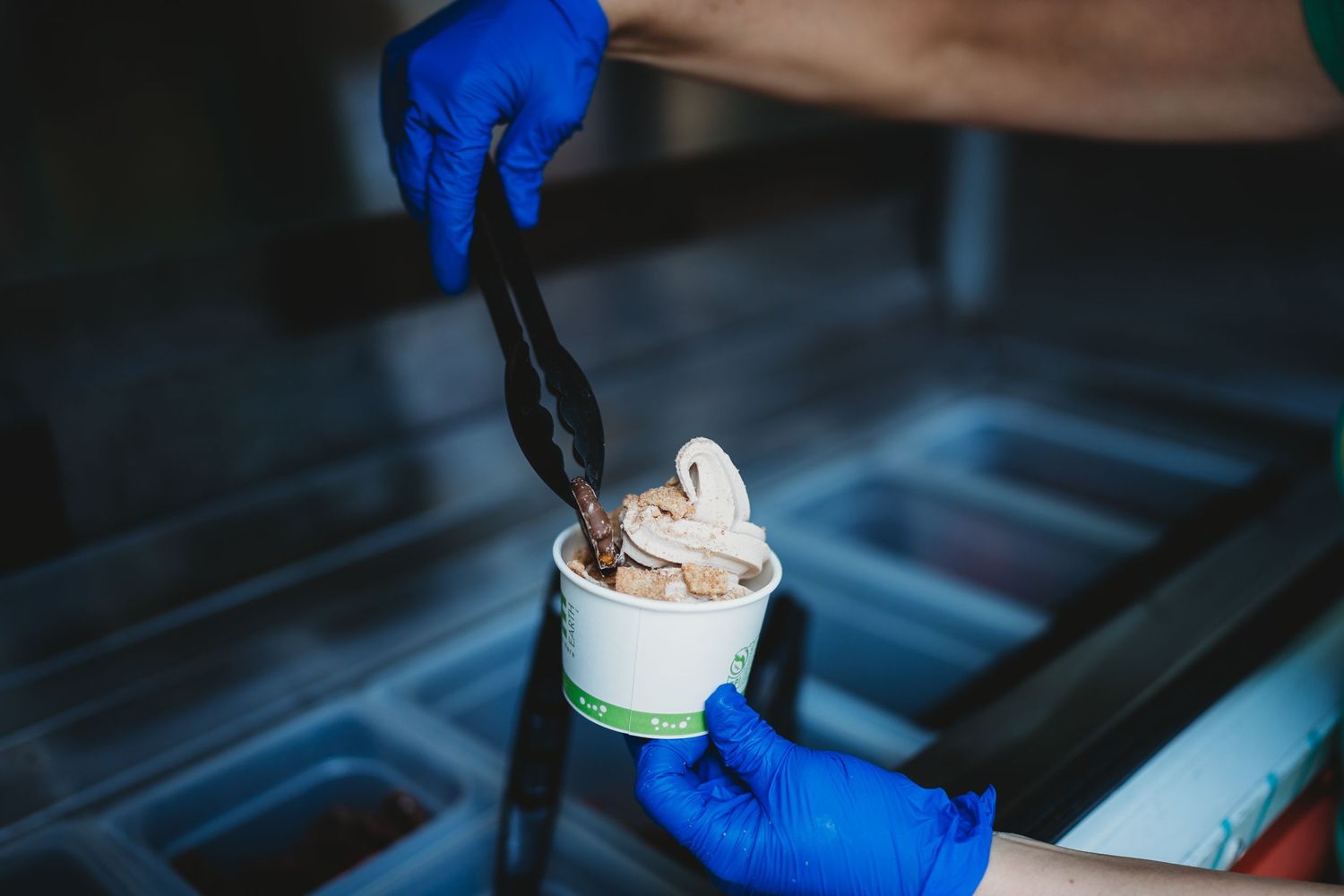 Soft serve ice cream being topped with dark sauce using blue gloved hands.