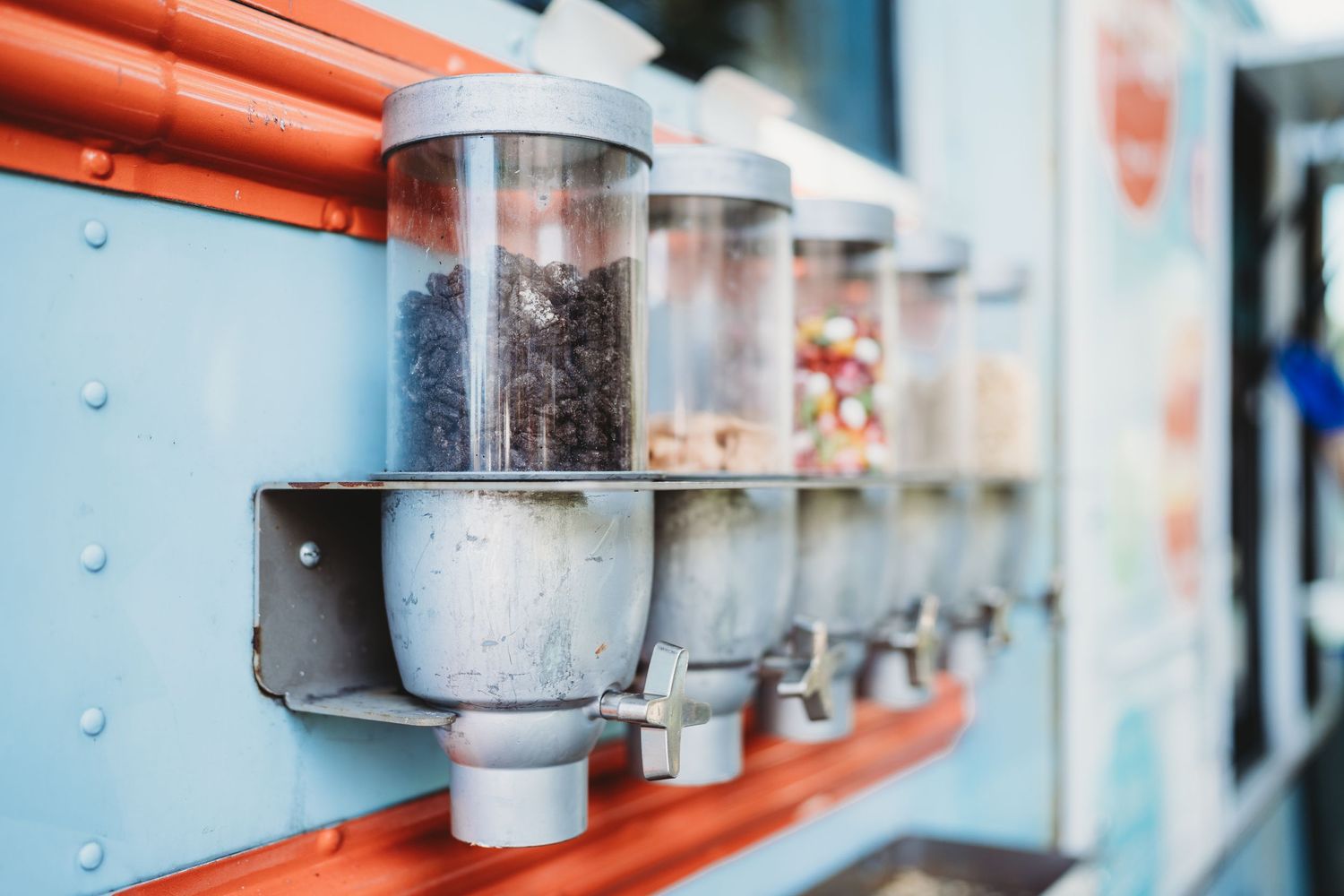 Row of transparent dispensers mounted on orange brackets containing various toppings.