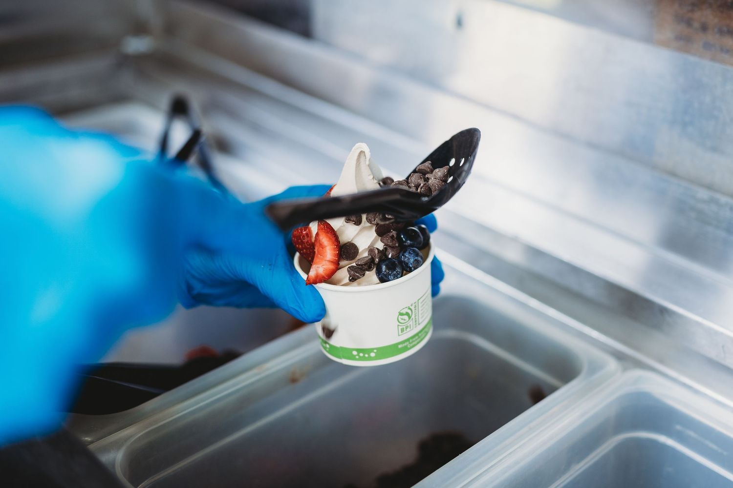 Hand in blue glove scooping toppings onto soft serve ice cream in a white cup.