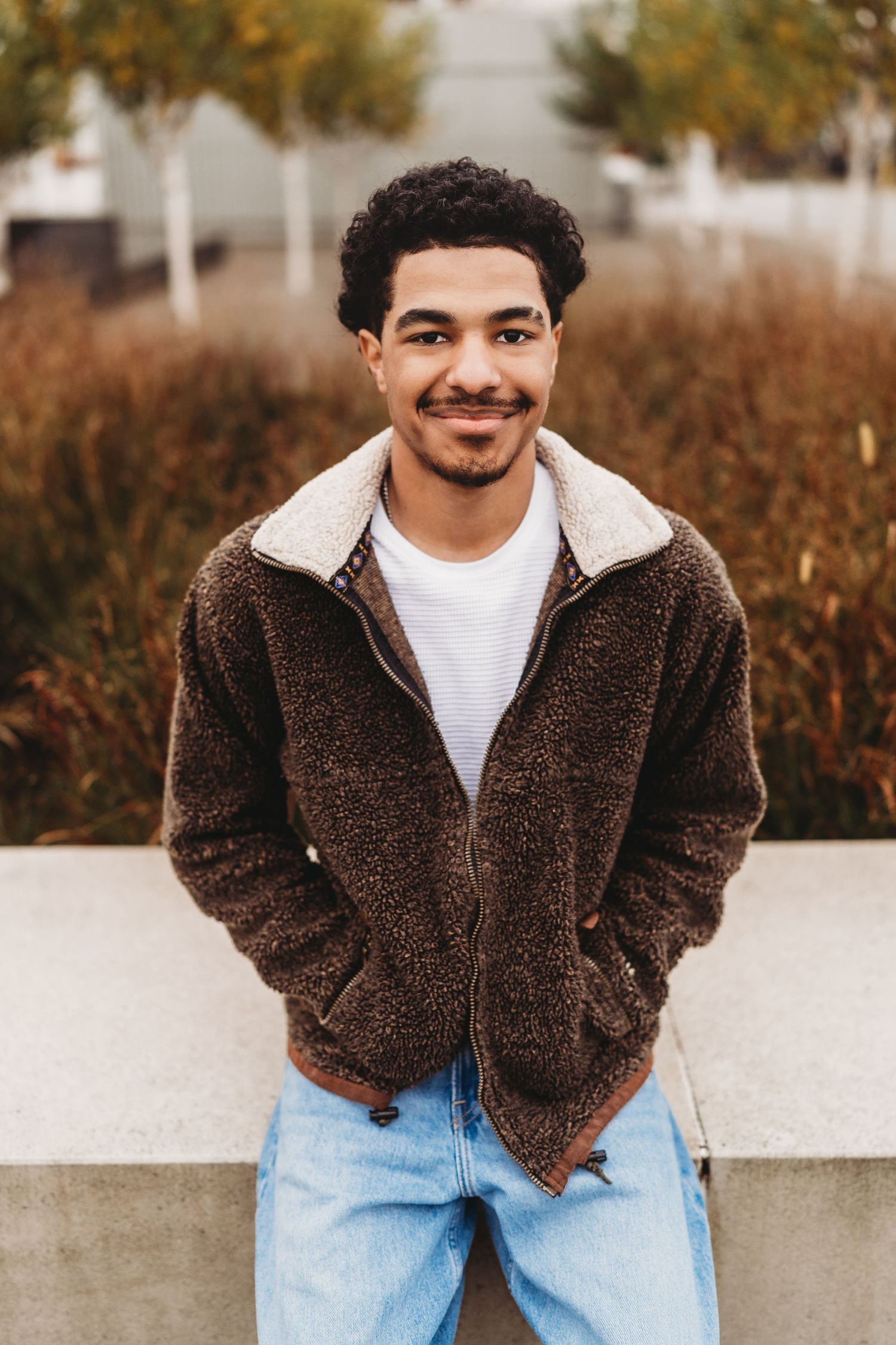 Young man in brown sherpa jacket and jeans smiling outdoors with autumn foliage in background.