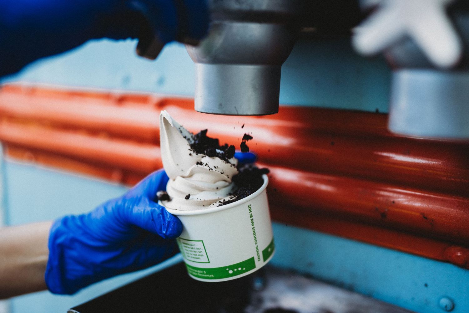 Ice cream being topped with chocolate sauce from a dispenser while wearing blue protective gloves.