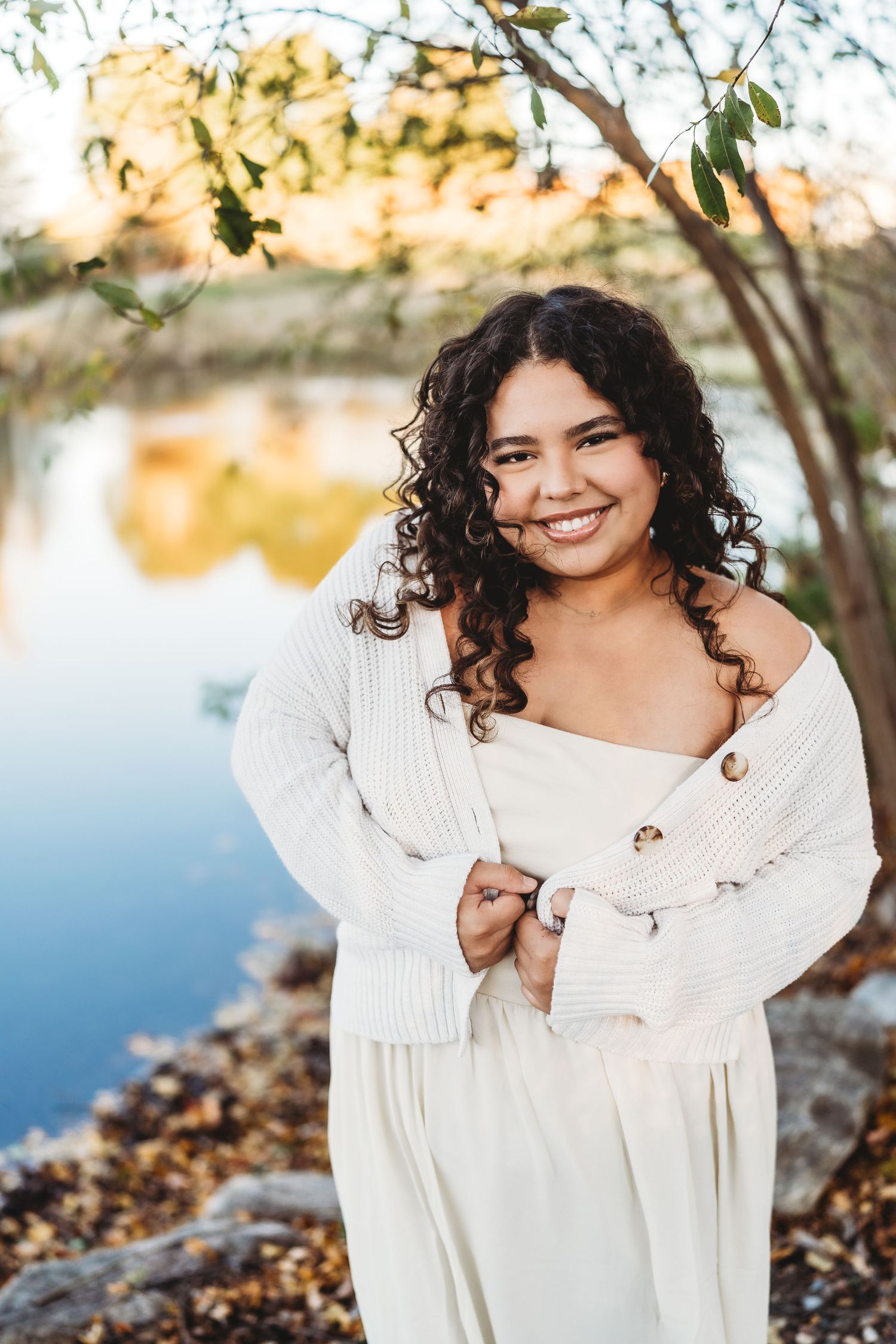 Smiling young woman with curly hair wearing a white dress and cardigan, posing outdoors near a reflective pond.