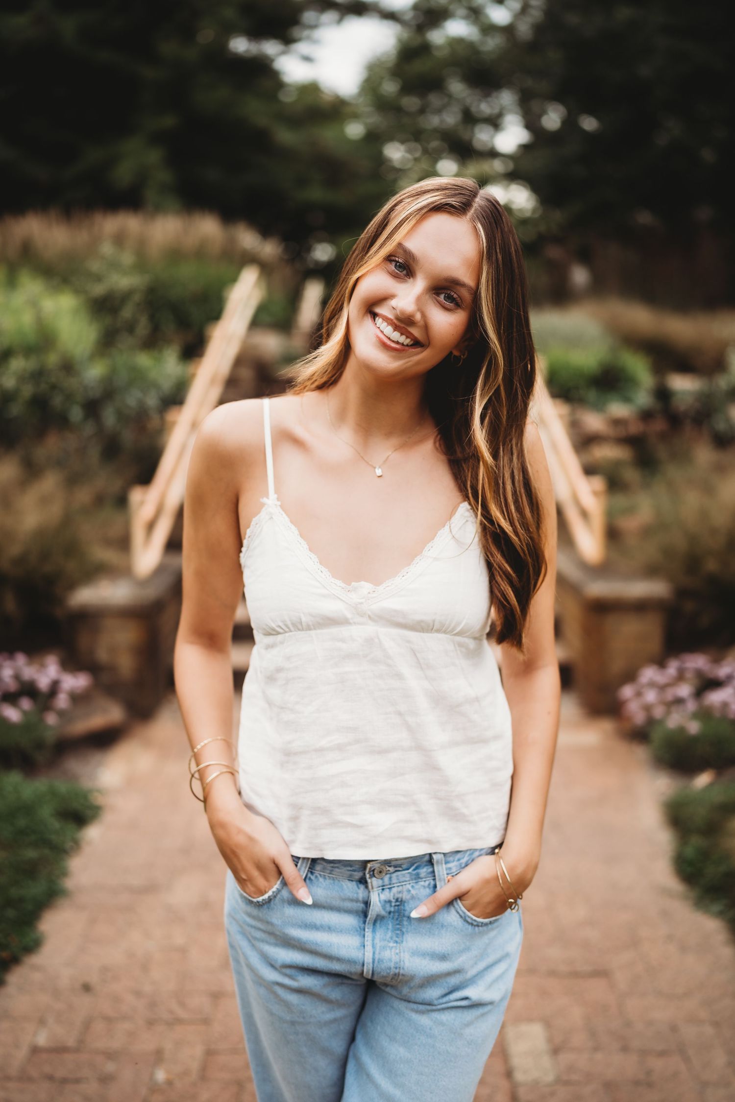 Smiling young woman in white camisole and light blue jeans posing outdoors in a garden setting.