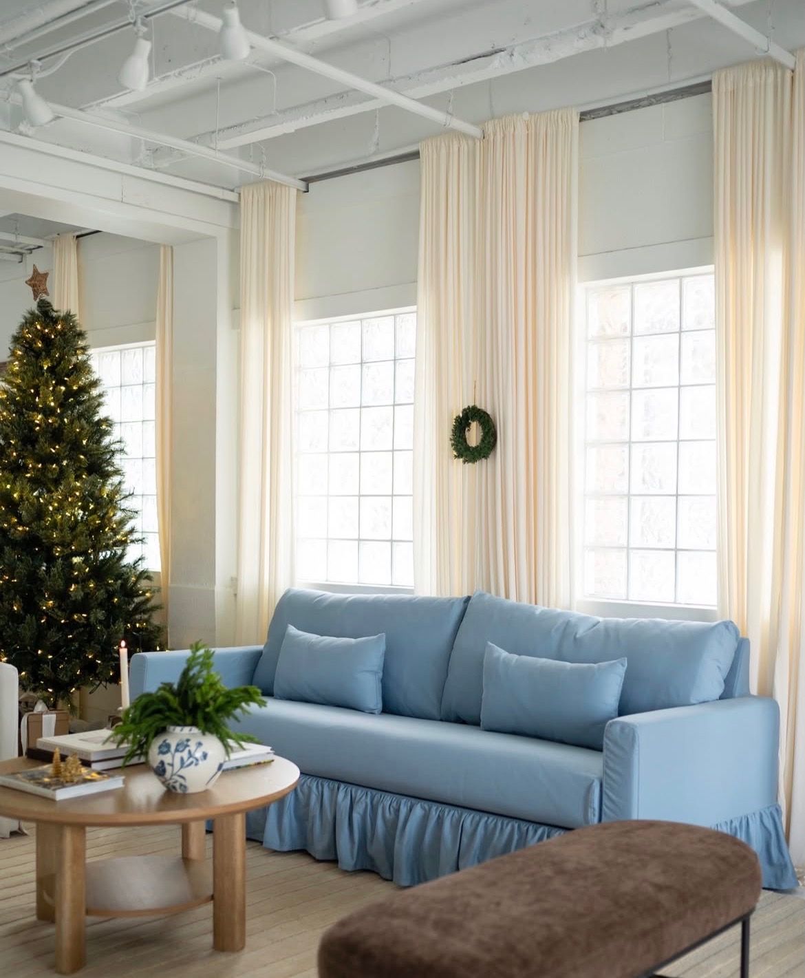 Cozy living room with a blue skirted sofa, cream curtains, a lit Christmas tree, round wooden coffee table and a wreath on tall windows.